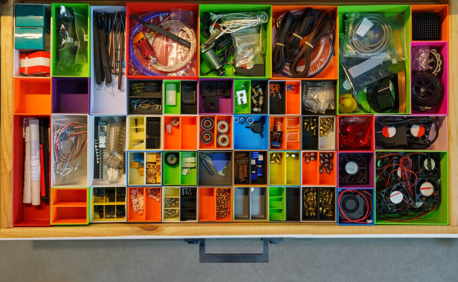 Gaveta de oficina vista de cima, cheia de organizadores modulares impressos em 3D em laranja, verde, roxo e azul, cada caixinha com parafusos, cabos e peças pequenas