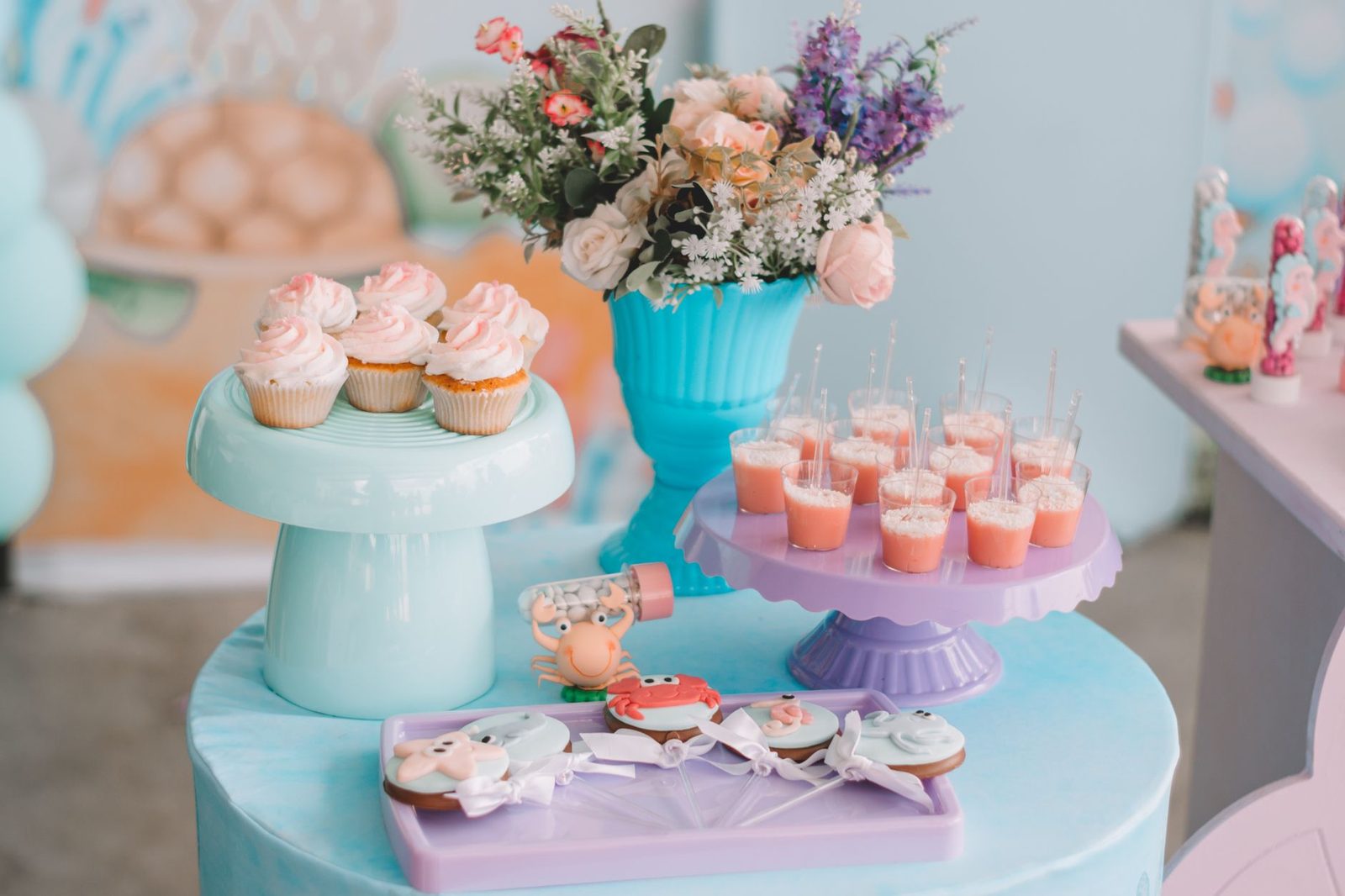 Mesa de festa infantil em tons pastel, com cupcakes em suporte azul-claro, copinhos de doce numa bandeja lilás e biscoitos decorados — o tipo de mesa onde a lembrancinha e a placa com nome entram
