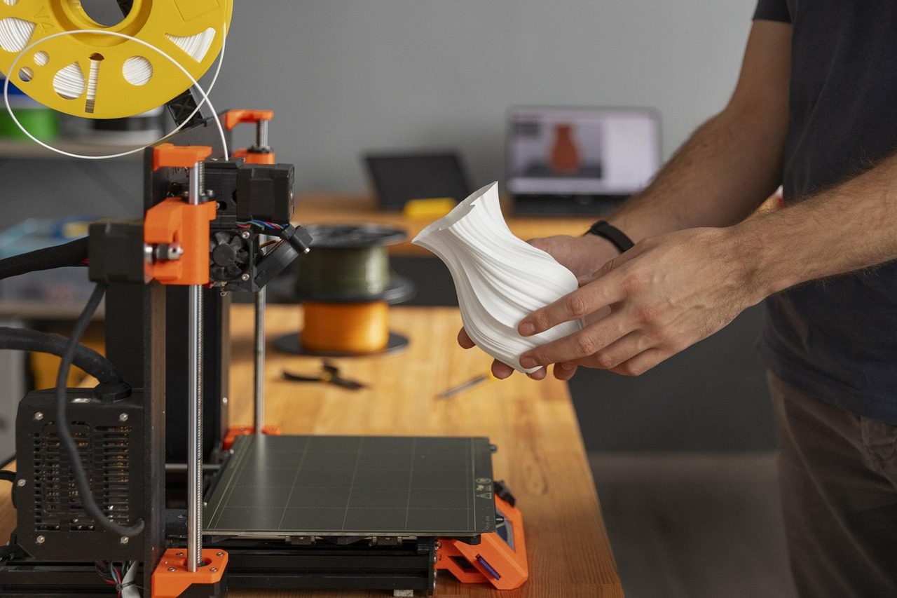 Pessoa segurando um vaso branco ao lado de uma impressora 3D com um rolo de filamento amarelo no topo
