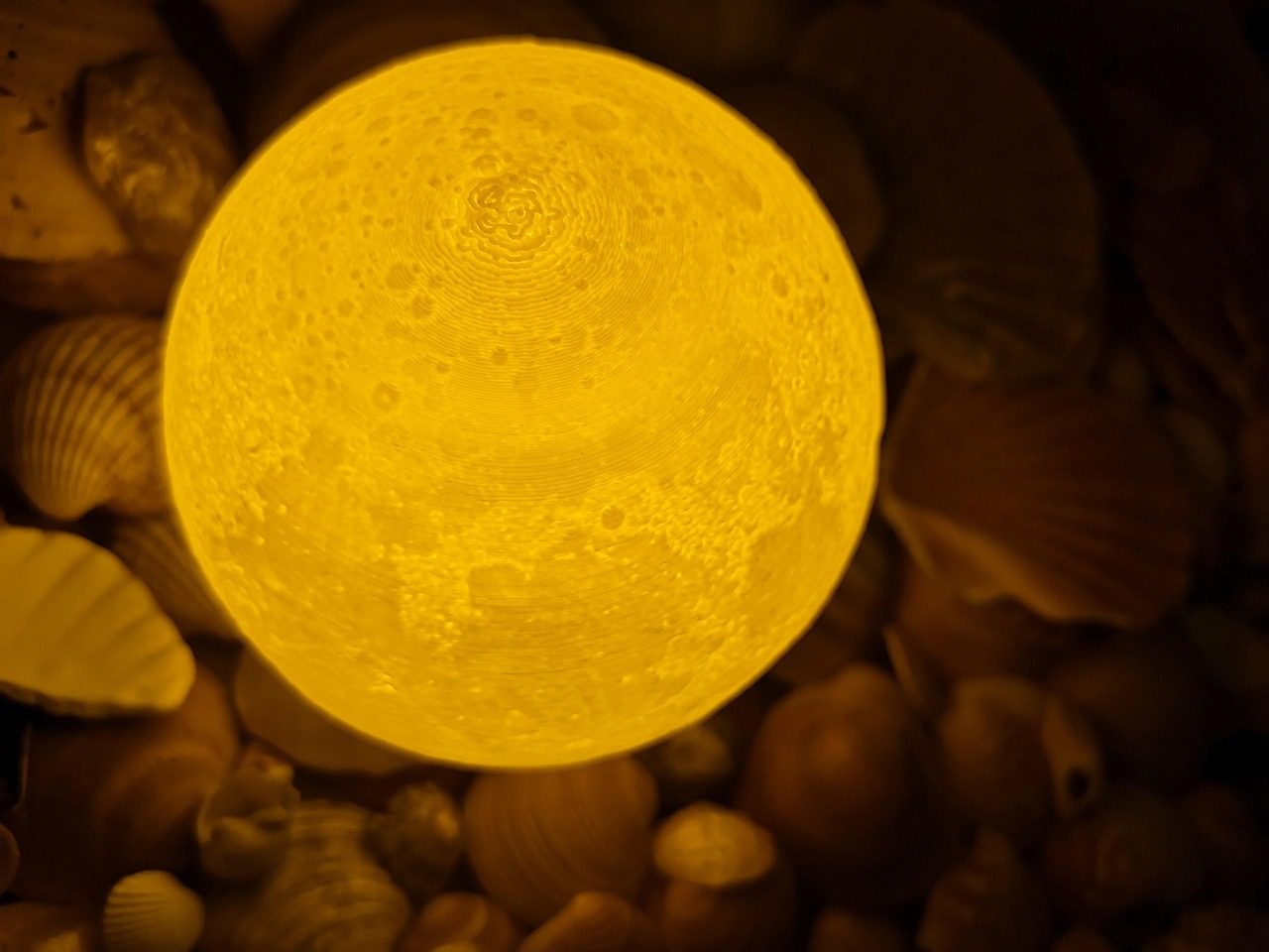 Luminária em forma de lua acesa: uma esfera amarela com relevo de crateras e anéis finos e concêntricos em volta do topo