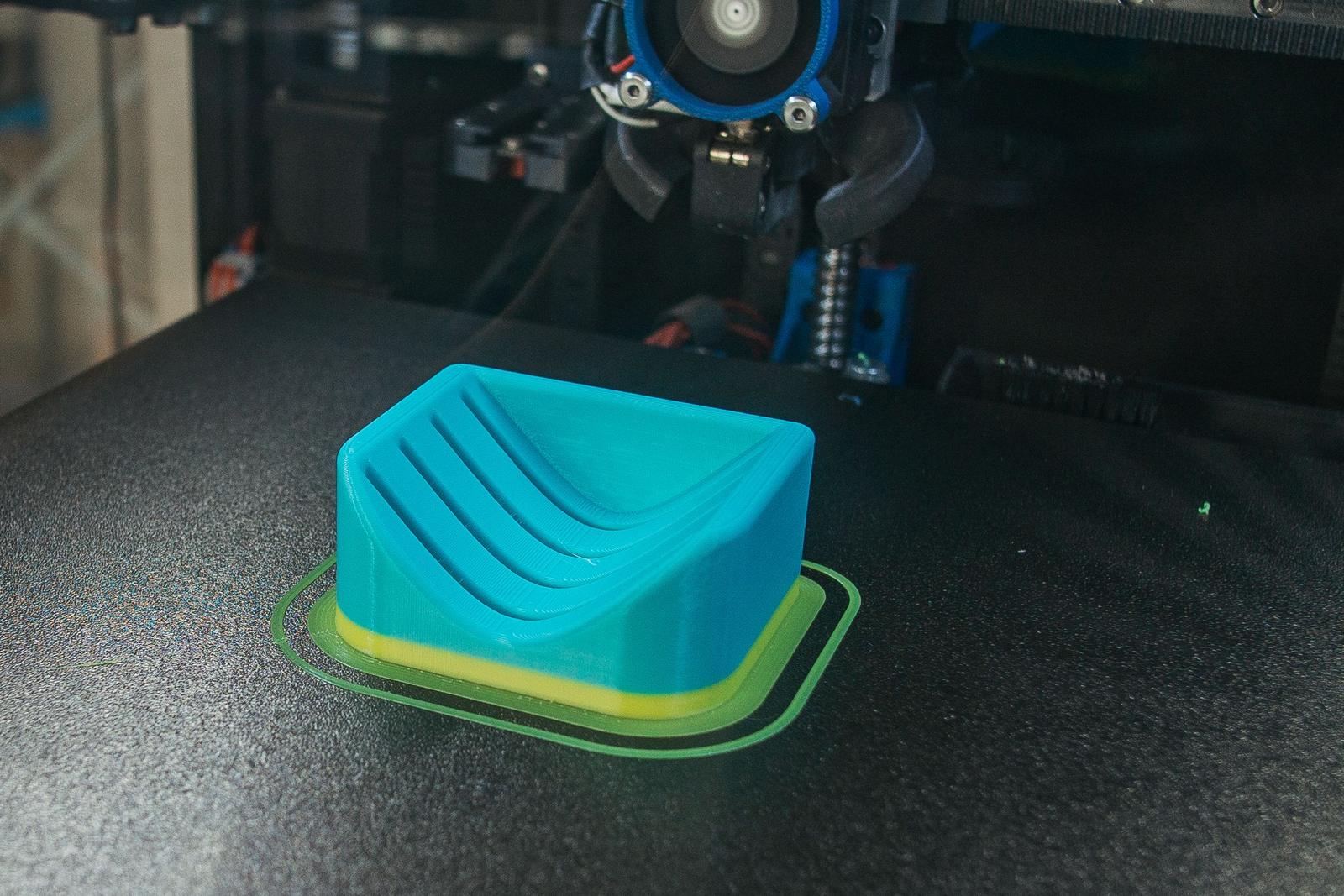 Peça de plástico azul com a face de cima curva e cheia de sulcos, base amarela, sobre a mesa de uma impressora com a cabeça de impressão logo acima