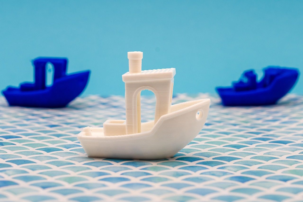 Benchy branco visto de lado, com casco curvo, cabine com janela em arco e chaminé redonda, e dois Benchys azuis desfocados ao fundo