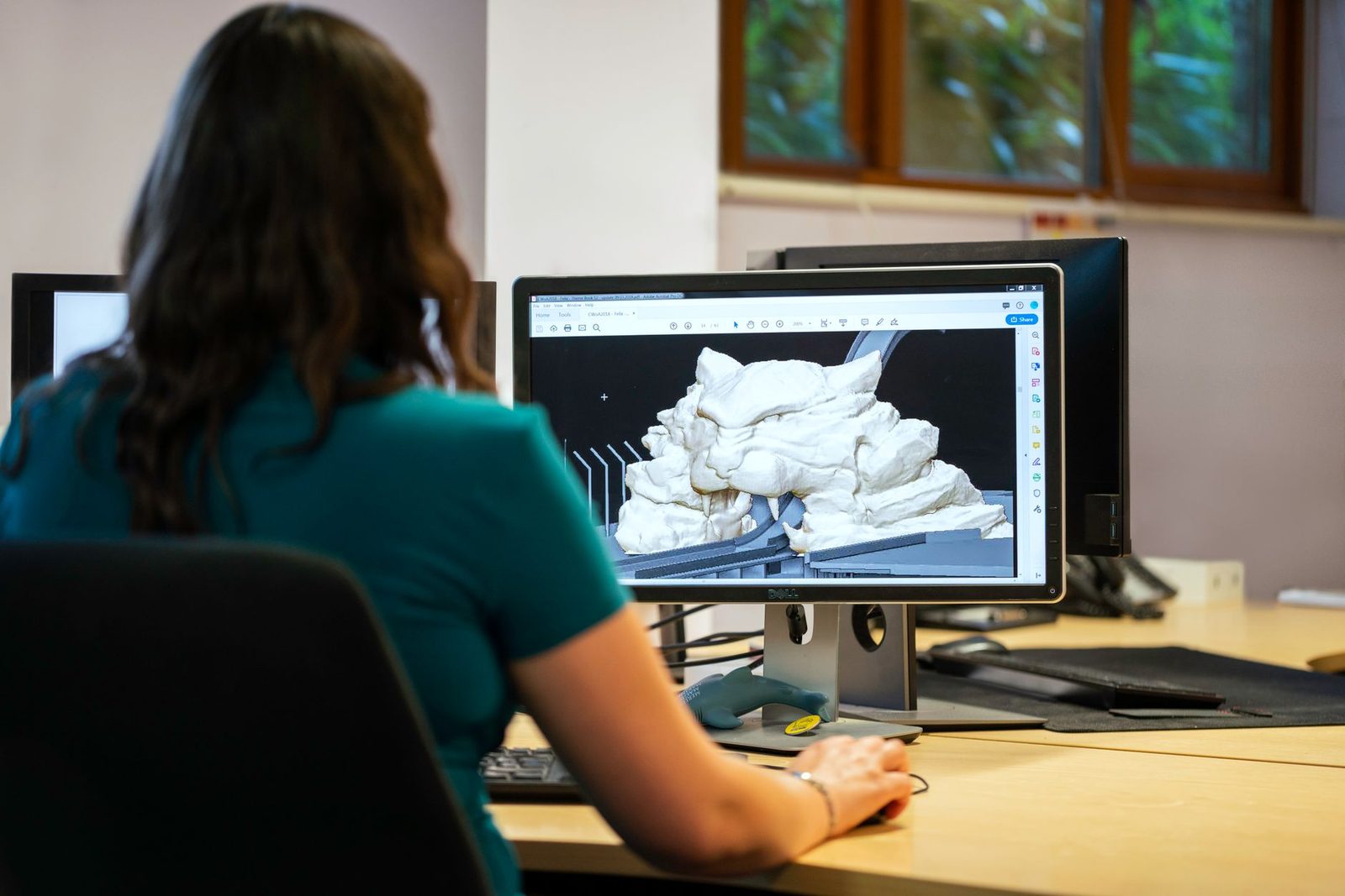 Mulher de costas diante de um monitor que mostra um modelo 3D esculpido, de forma orgânica e cor clara