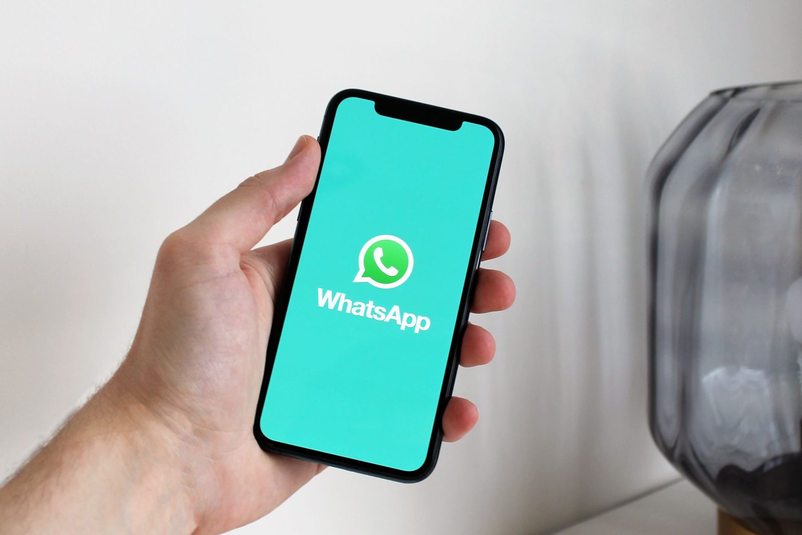 Mão segurando um celular com o logo do WhatsApp na tela, sobre fundo claro