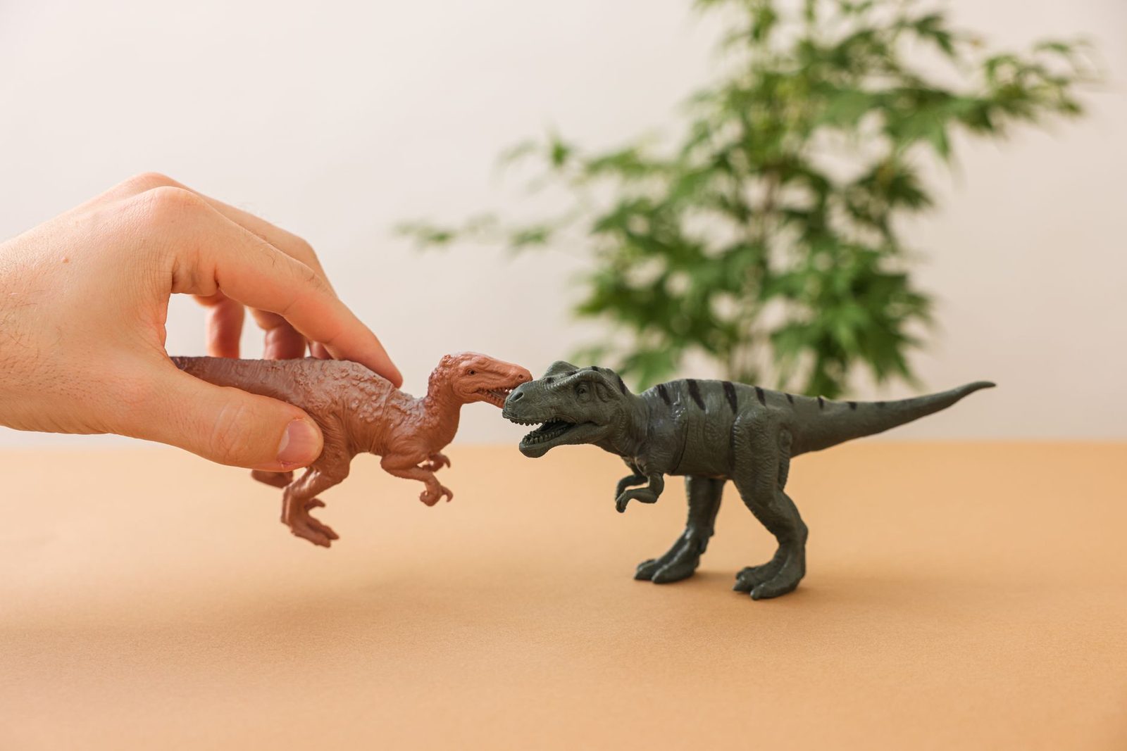 Mão segurando um dinossauro de brinquedo marrom de frente pra um T-Rex de brinquedo cinza, sobre mesa bege com uma planta desfocada ao fundo