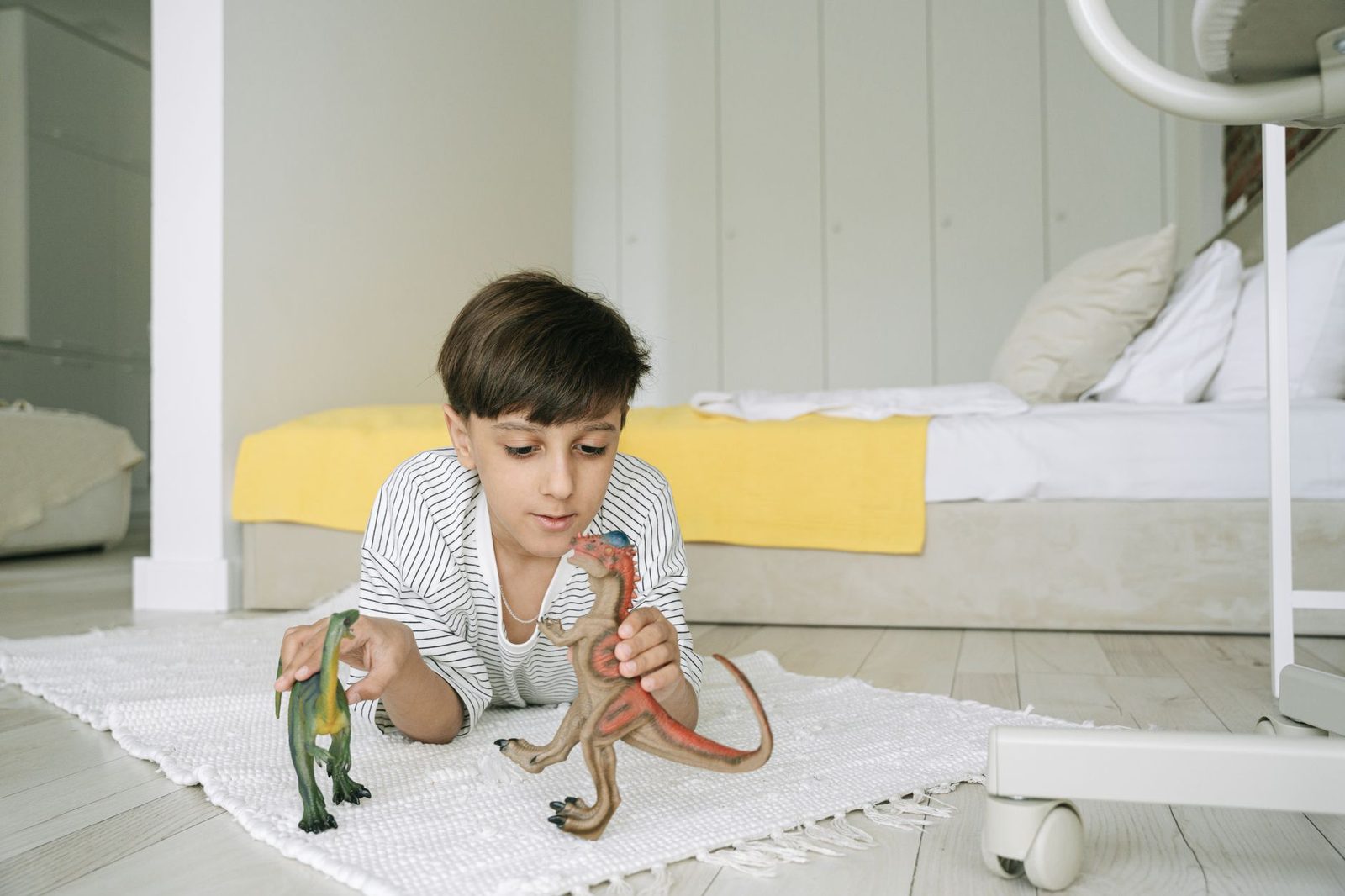 Menino deitado de bruços num tapete branco brincando com dois dinossauros de brinquedo, um verde e um marrom, num quarto claro com cama ao fundo