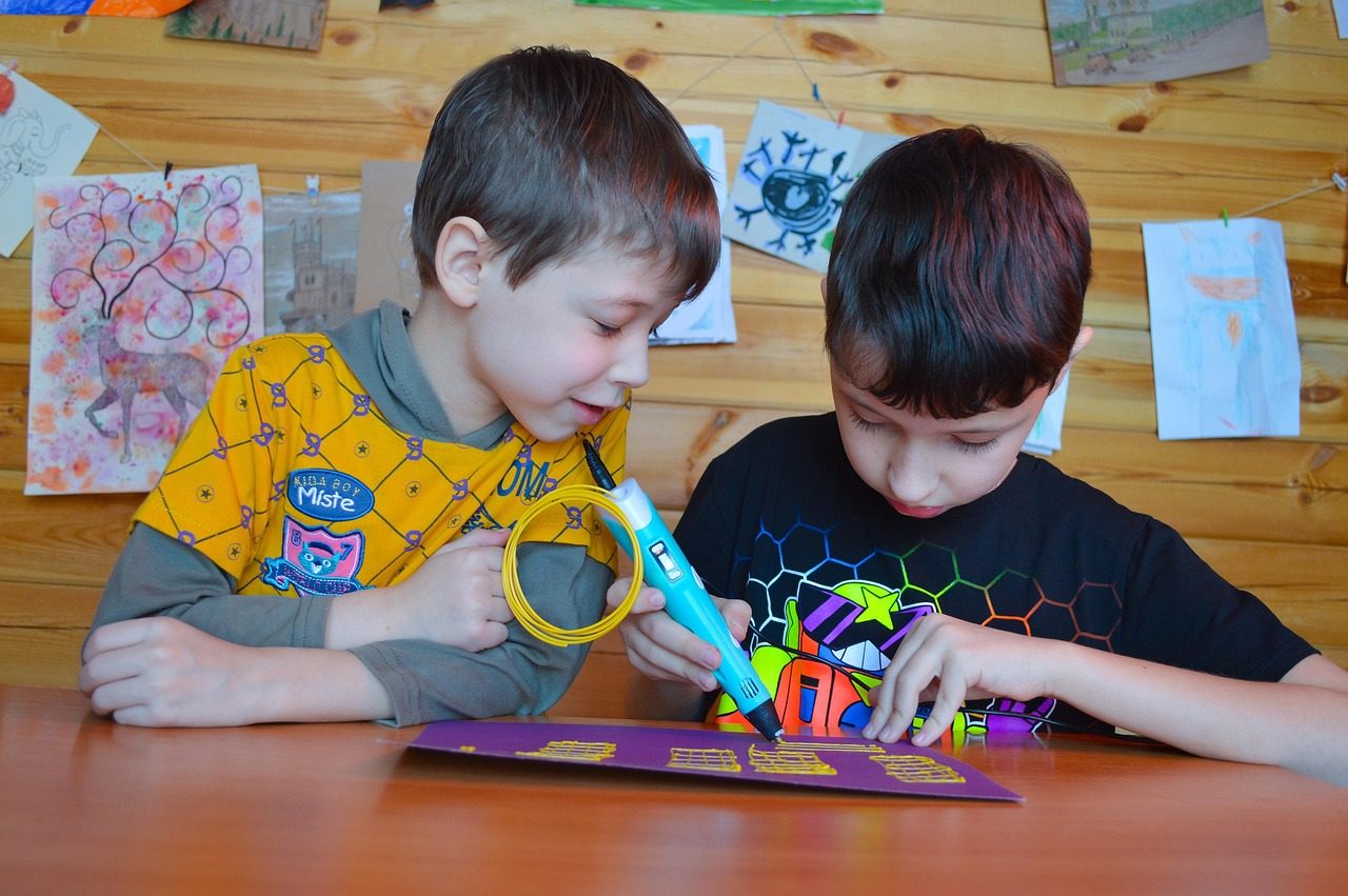 Dois meninos à mesa: um desenha com uma caneta 3D azul e filamento amarelo sobre uma folha roxa, onde já há várias peças planas em grade feitas com o plástico amarelo