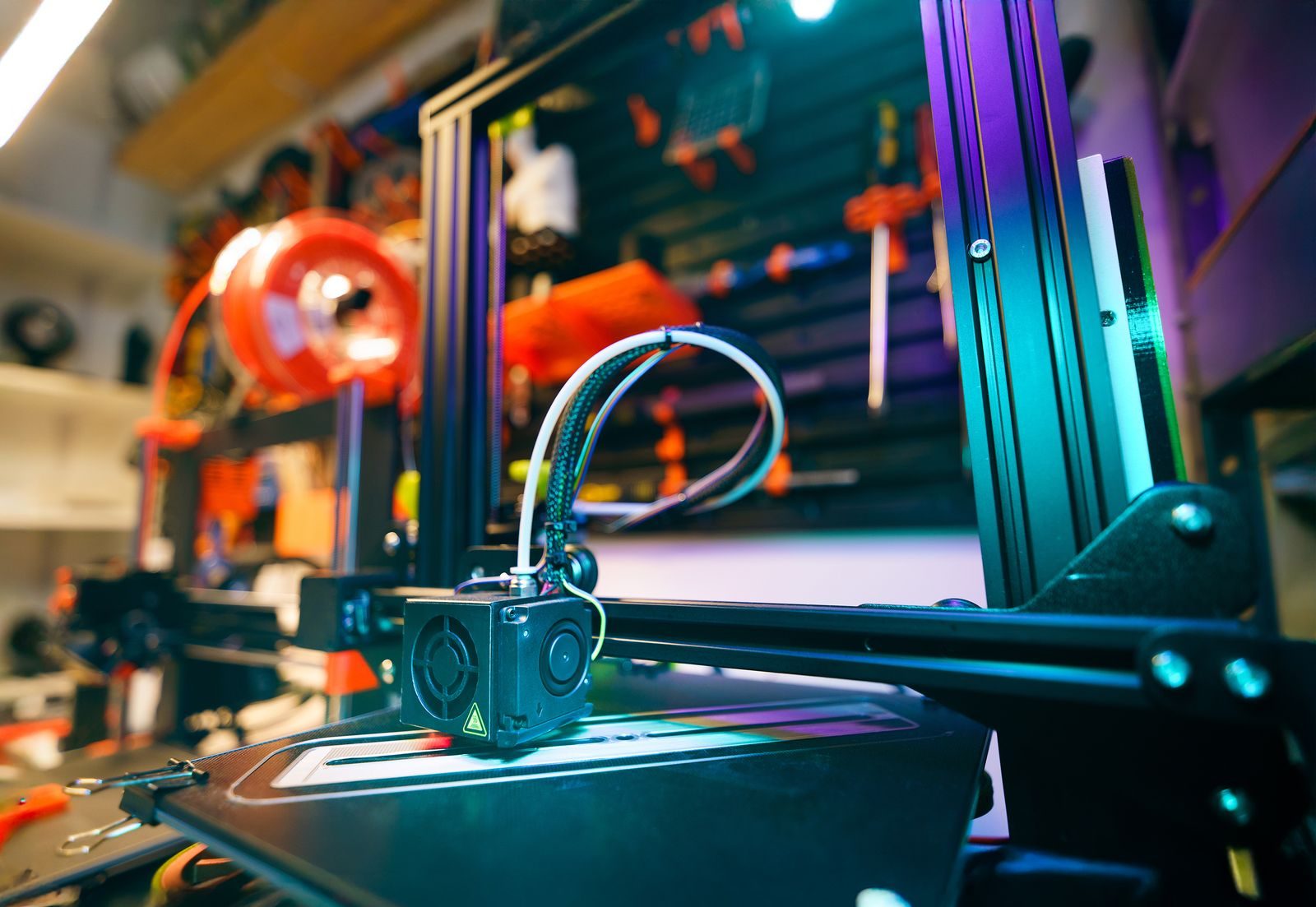 Cabeçote de uma impressora 3D de filamento parado sobre a mesa, numa oficina com carretel laranja ao fundo