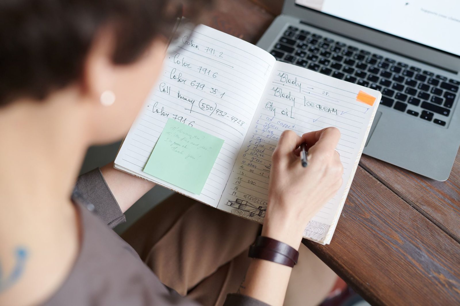 Pessoa anotando à mão num caderno aberto, com um notebook ligado ao lado sobre a mesa de madeira
