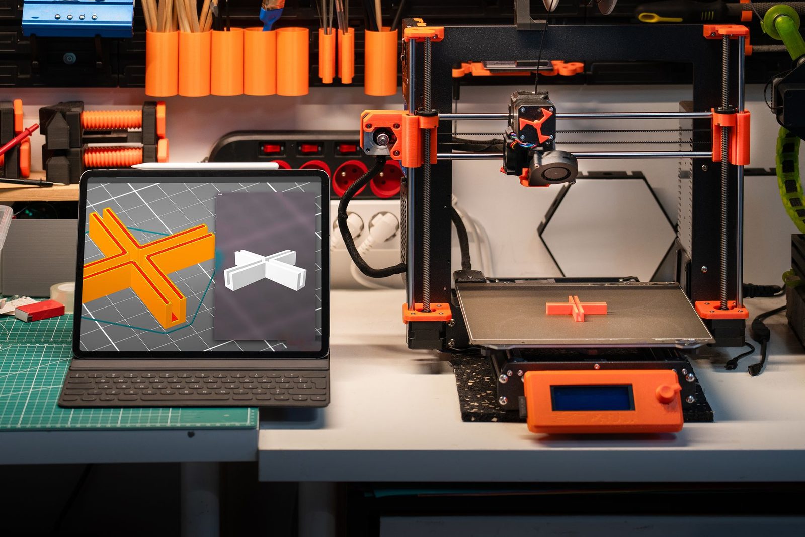 Tablet na bancada com o fatiador aberto mostrando uma peça em cruz laranja já fatiada, ao lado de uma impressora 3D com a mesma peça impressa na mesa
