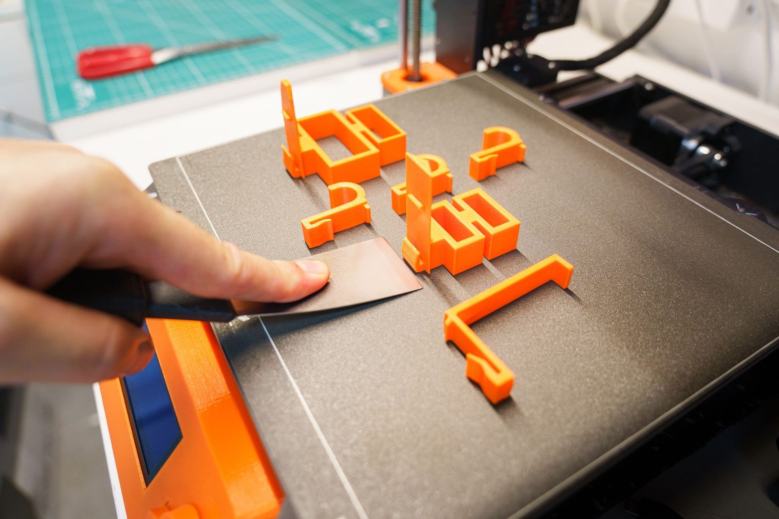 Mão usando uma espátula pra soltar peças pequenas de PLA laranja da mesa PEI texturizada de uma impressora 3D, com o tapete de corte verde ao fundo