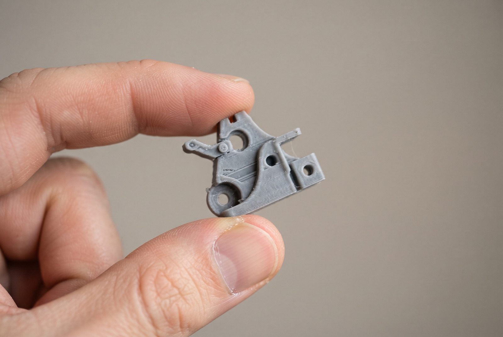 Peça pequena impressa em 3D segurada entre os dedos contra um fundo neutro, mostrando o tamanho real
