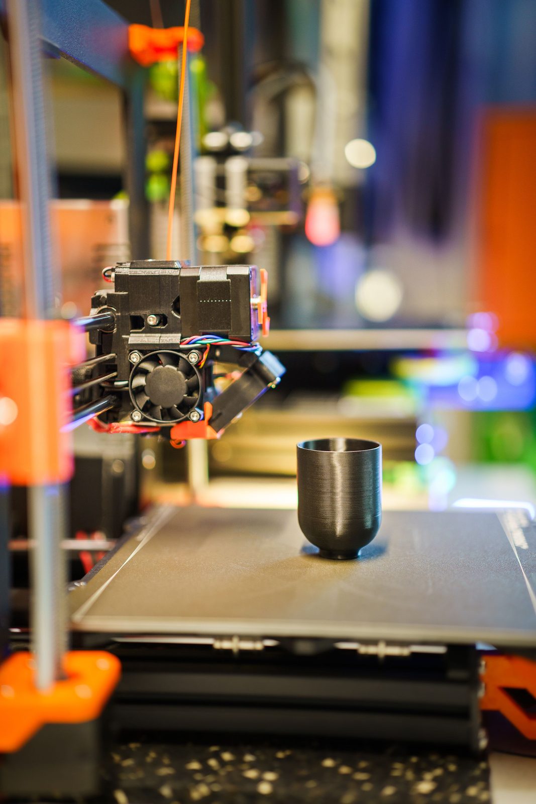 Copo preto de plástico com finas linhas horizontais na parede, em pé na mesa de uma impressora 3D de filamento, com o cabeçote ao lado