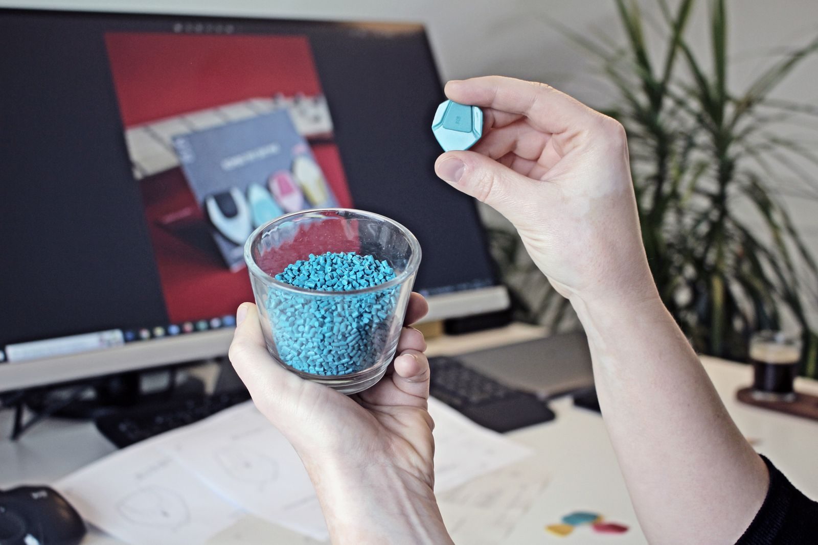 Mãos segurando um copo de vidro cheio de grãos de plástico azul e uma peça azul pronta, na frente de um monitor