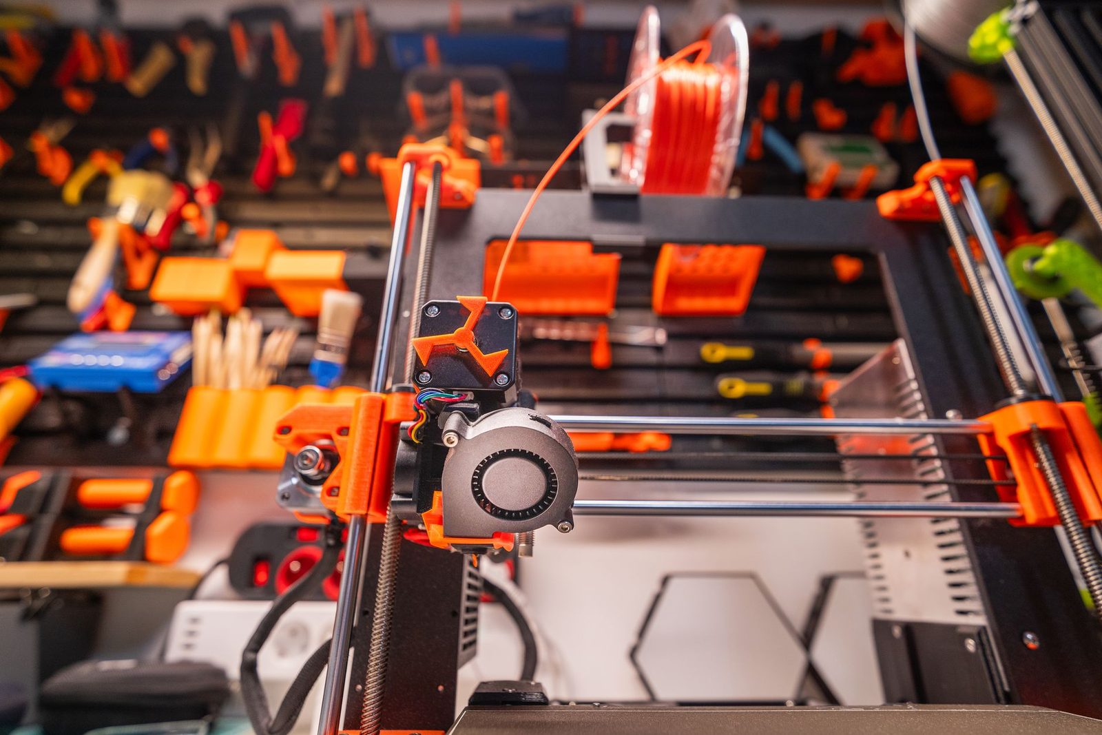 Cabeça de impressão de uma impressora 3D FDM, com ventoinha e cabos à mostra, diante de um painel de ferramentas na oficina