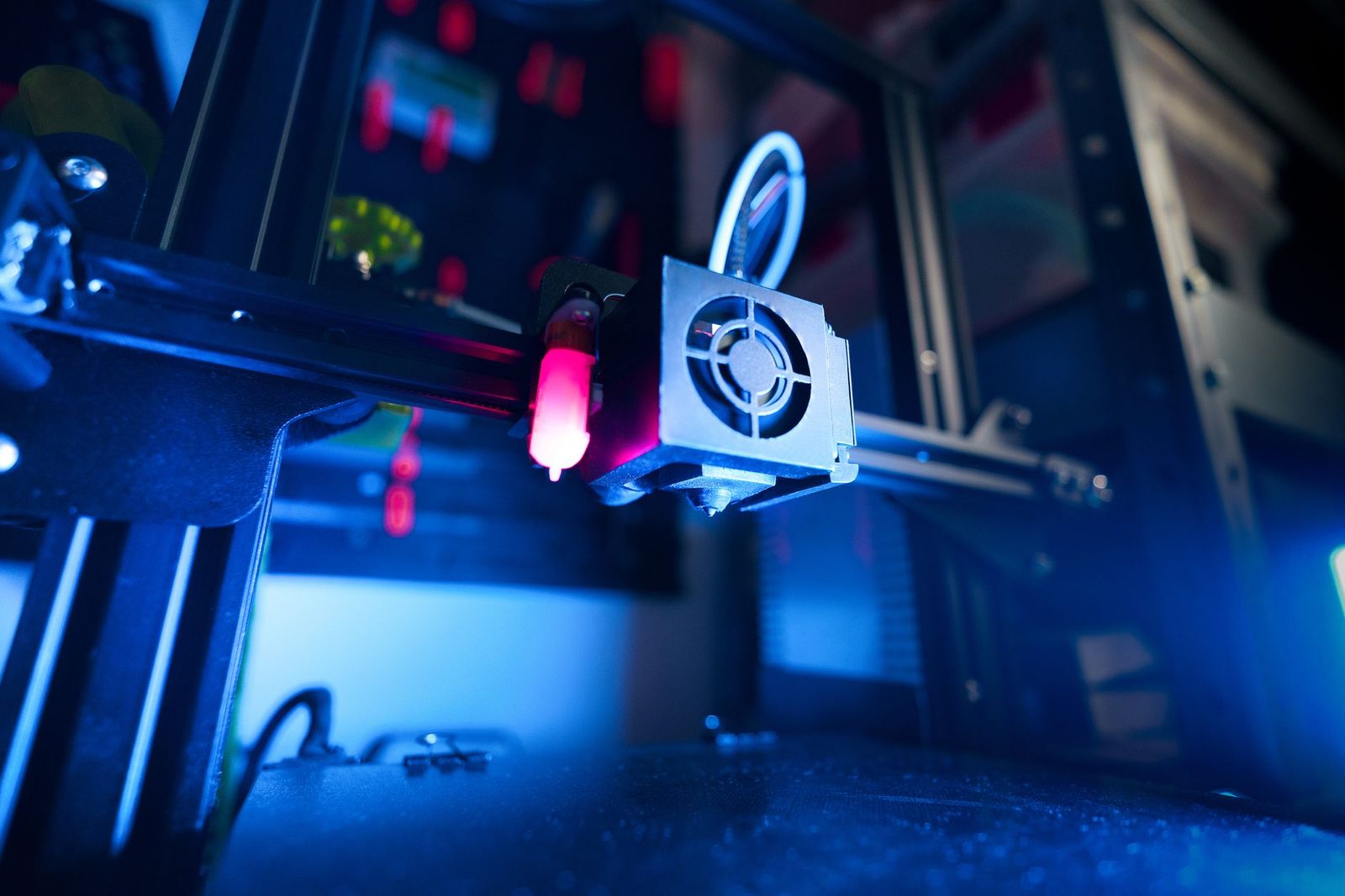 Cabeça de impressão de uma impressora 3D sob luz azul, com o tubo de teflon branco fazendo uma curva e entrando por cima