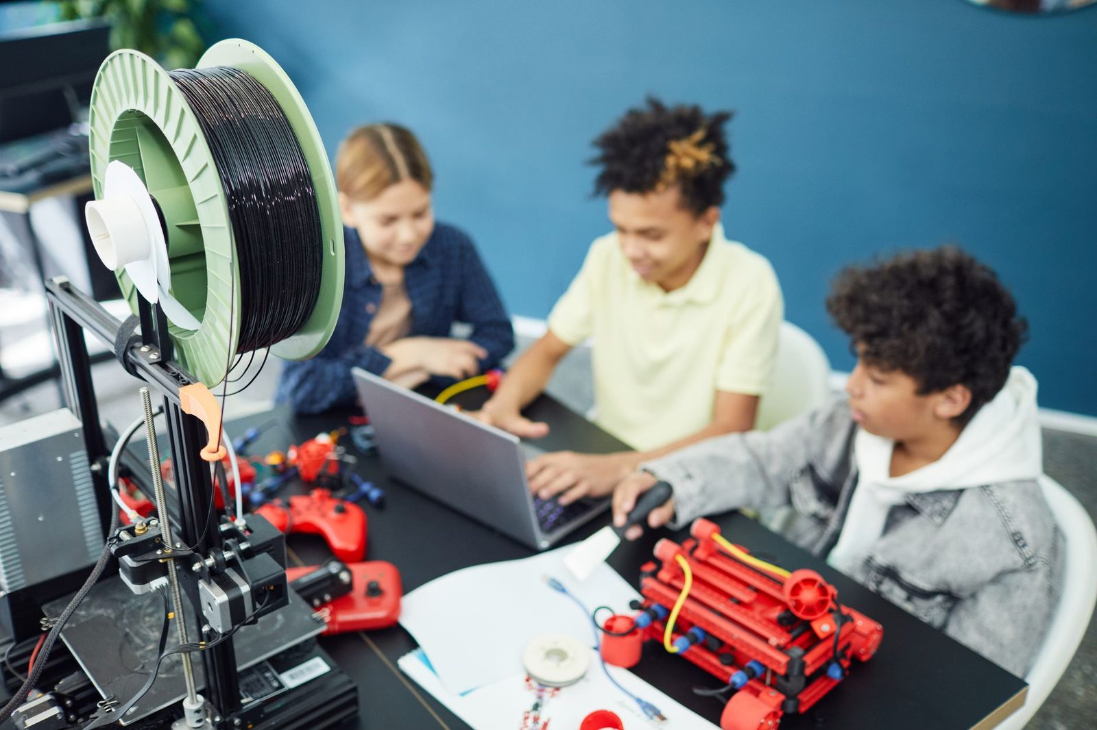 Três estudantes numa mesa de laboratório de robótica, com notebook e peças de robô vermelhas, ao lado de uma impressora 3D com carretel de filamento preto