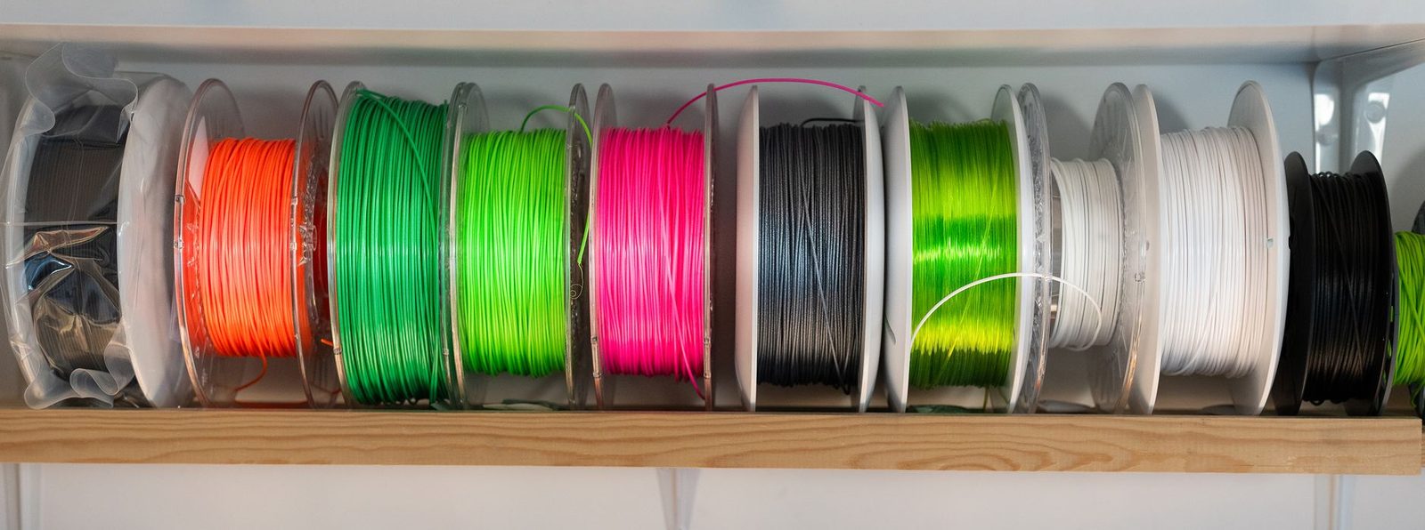 Fileira de rolos de filamento em cores diferentes numa prateleira de madeira: laranja, dois tons de verde, rosa choque, cinza escuro, verde translúcido, branco e preto