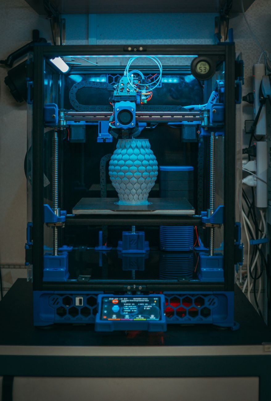 Vaso com textura de escamas sendo impresso dentro de uma impressora 3D fechada, iluminada por luz azul