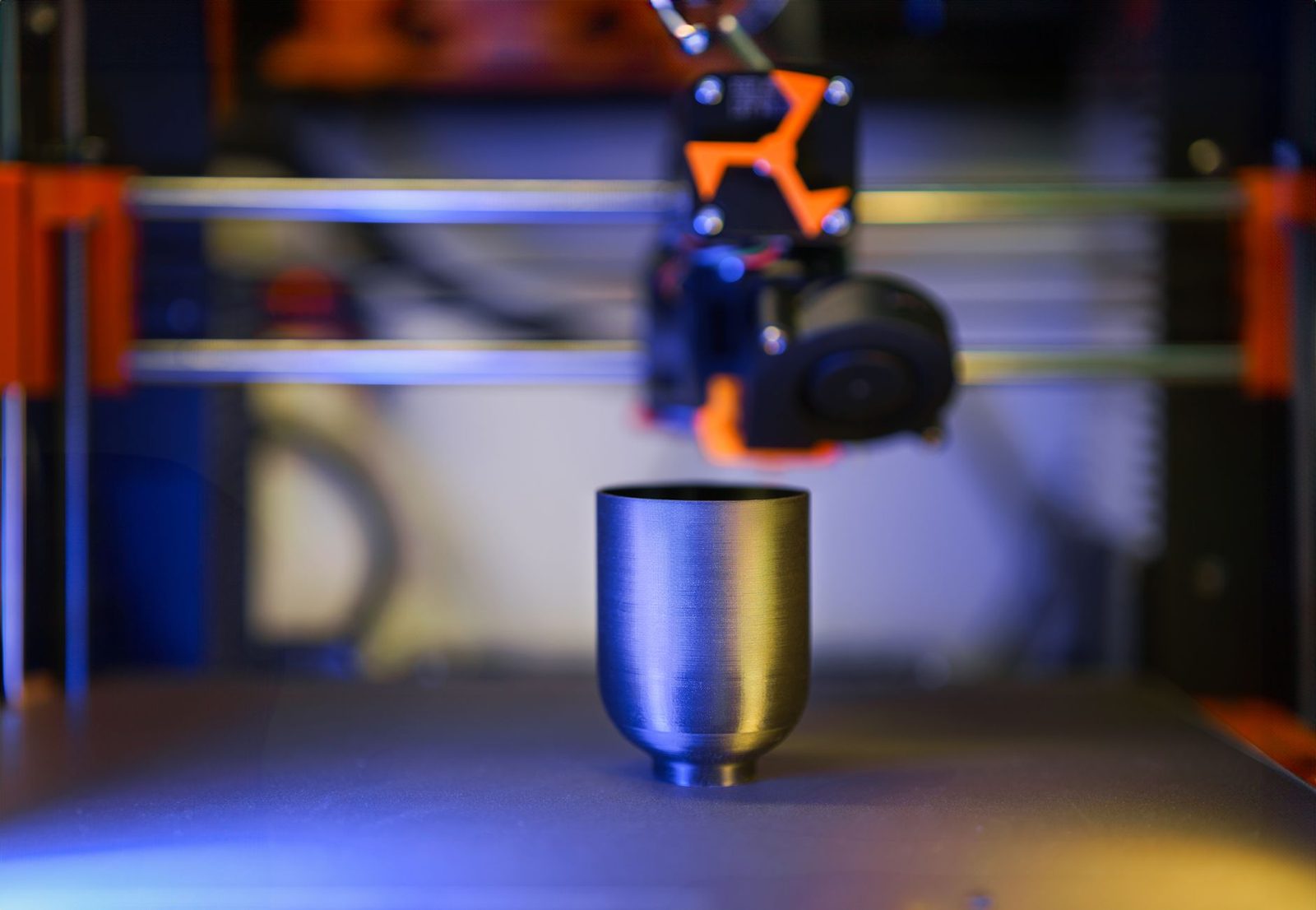 Copo impresso em 3D com filamento brilhante metálico sobre a mesa da impressora, com as linhas de camada aparecendo no reflexo da luz