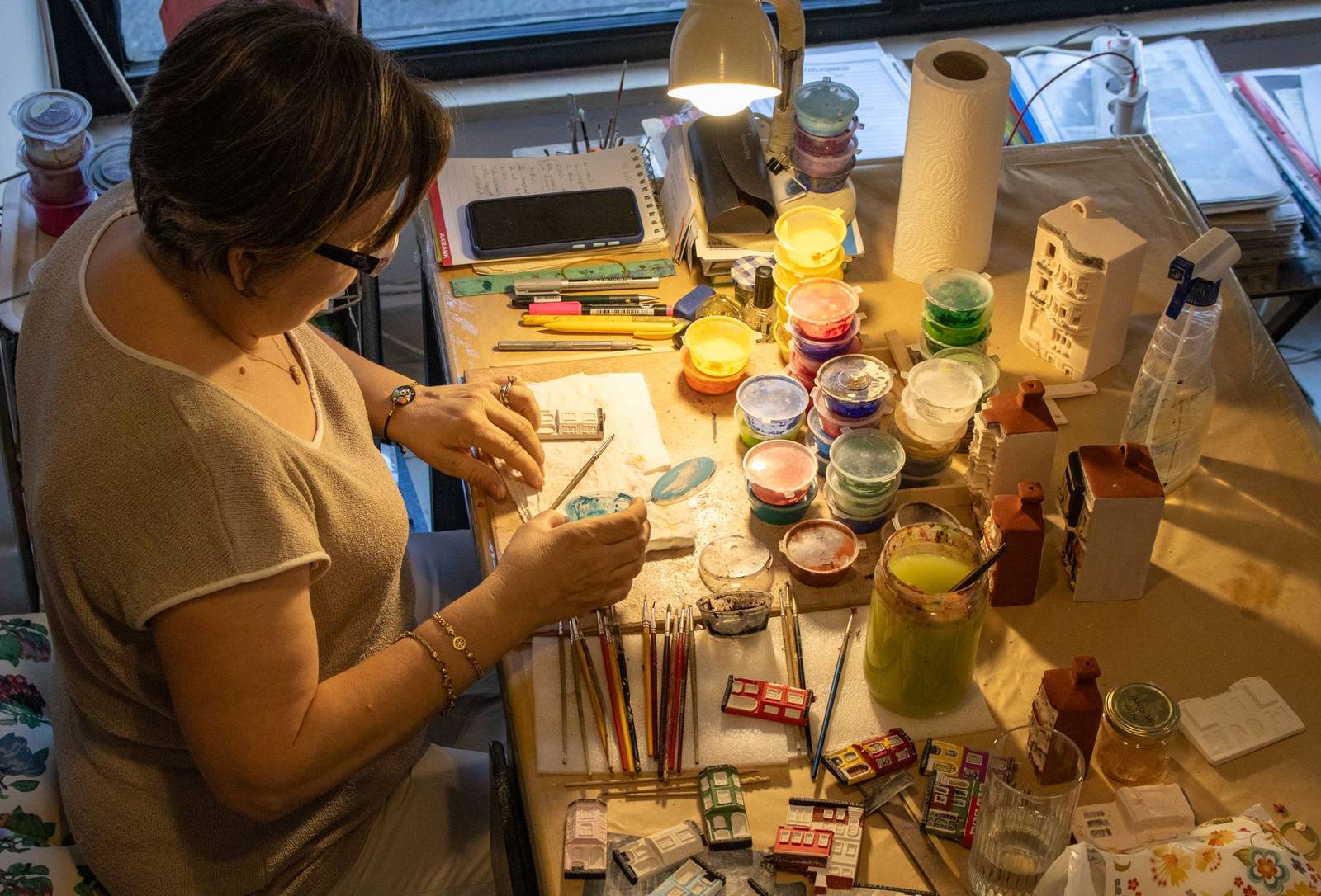 Artesã pintando com pincel miniaturas de casinhas numa bancada cheia de potes de tinta