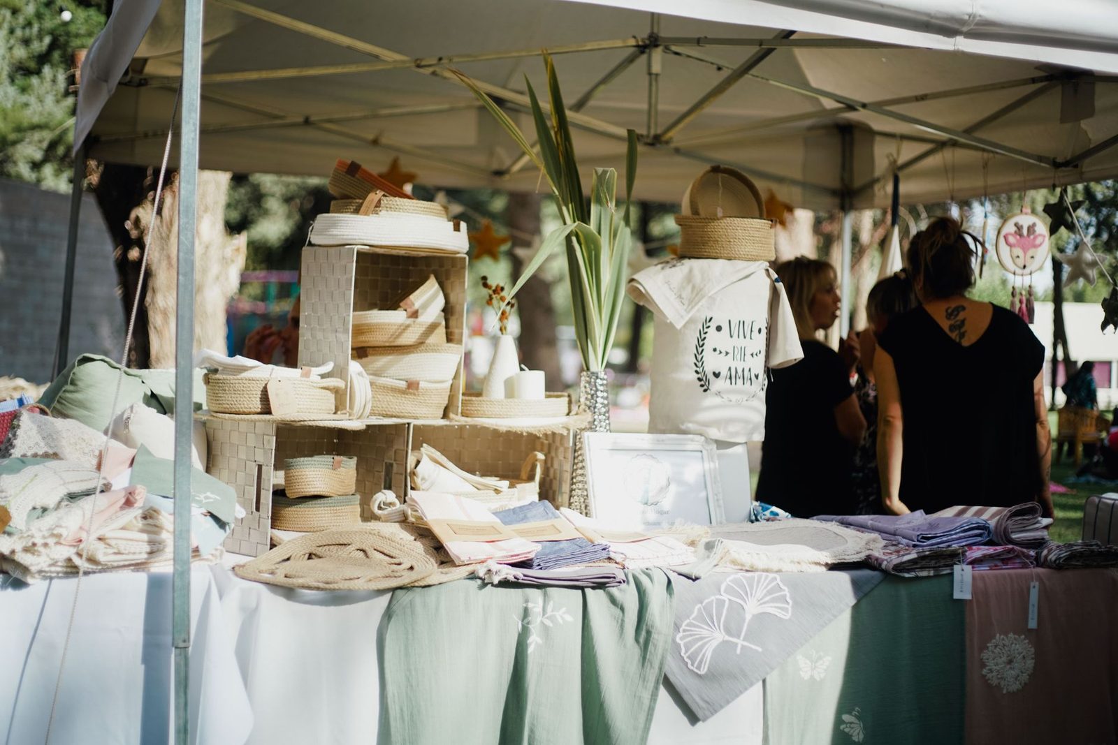 Banca de feira de artesanato sob uma tenda, com cestos de corda, panos bordados e etiquetas penduradas nos produtos