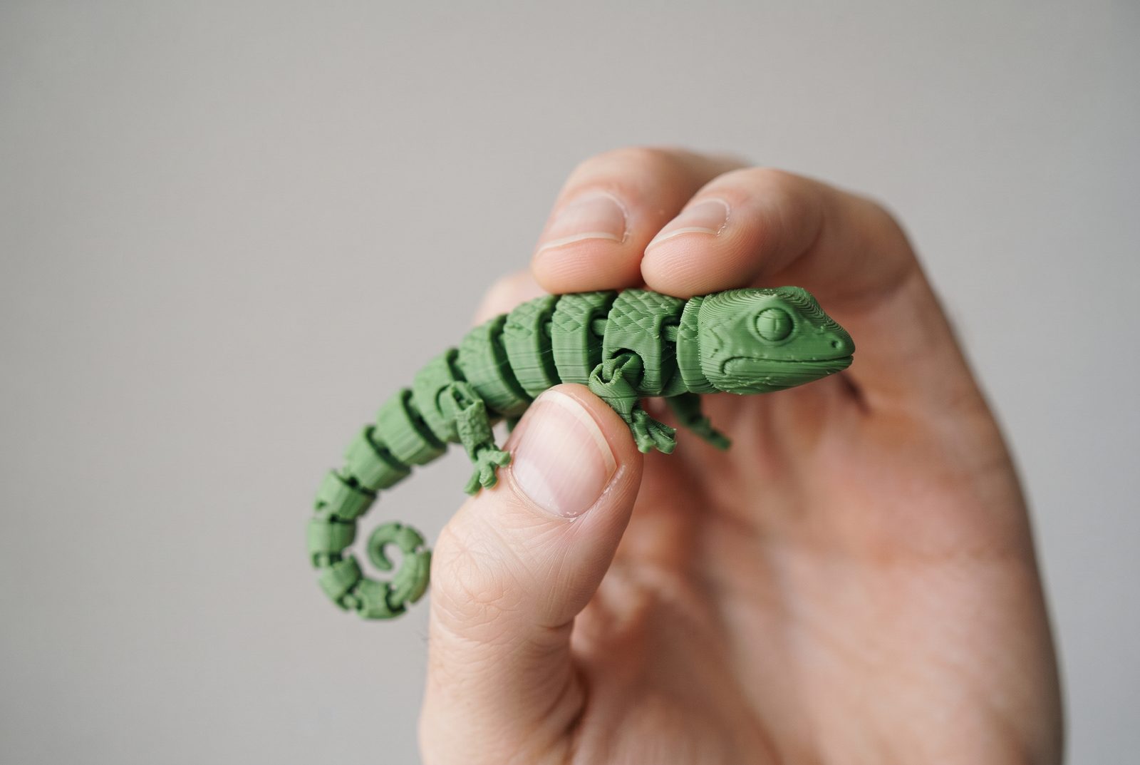 Mão segurando um lagarto articulado impresso em 3D em PLA verde sobre fundo claro liso