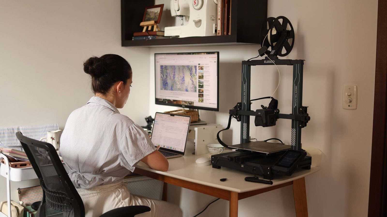 Mulher trabalhando no notebook numa escrivaninha em casa, com uma impressora 3D de filamento logo ao lado do monitor