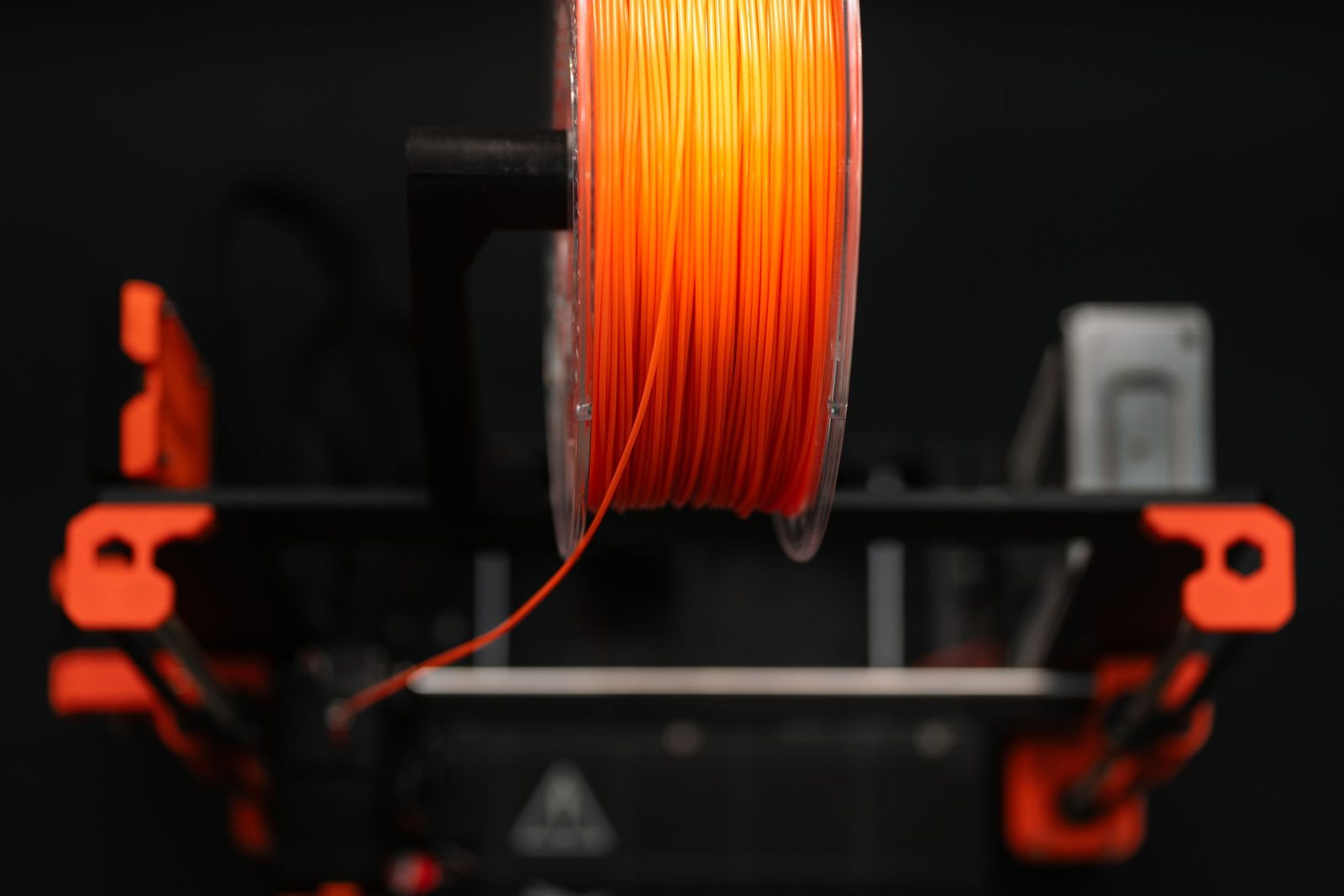 Carretel de plástico transparente com filamento laranja num suporte no alto de uma impressora 3D