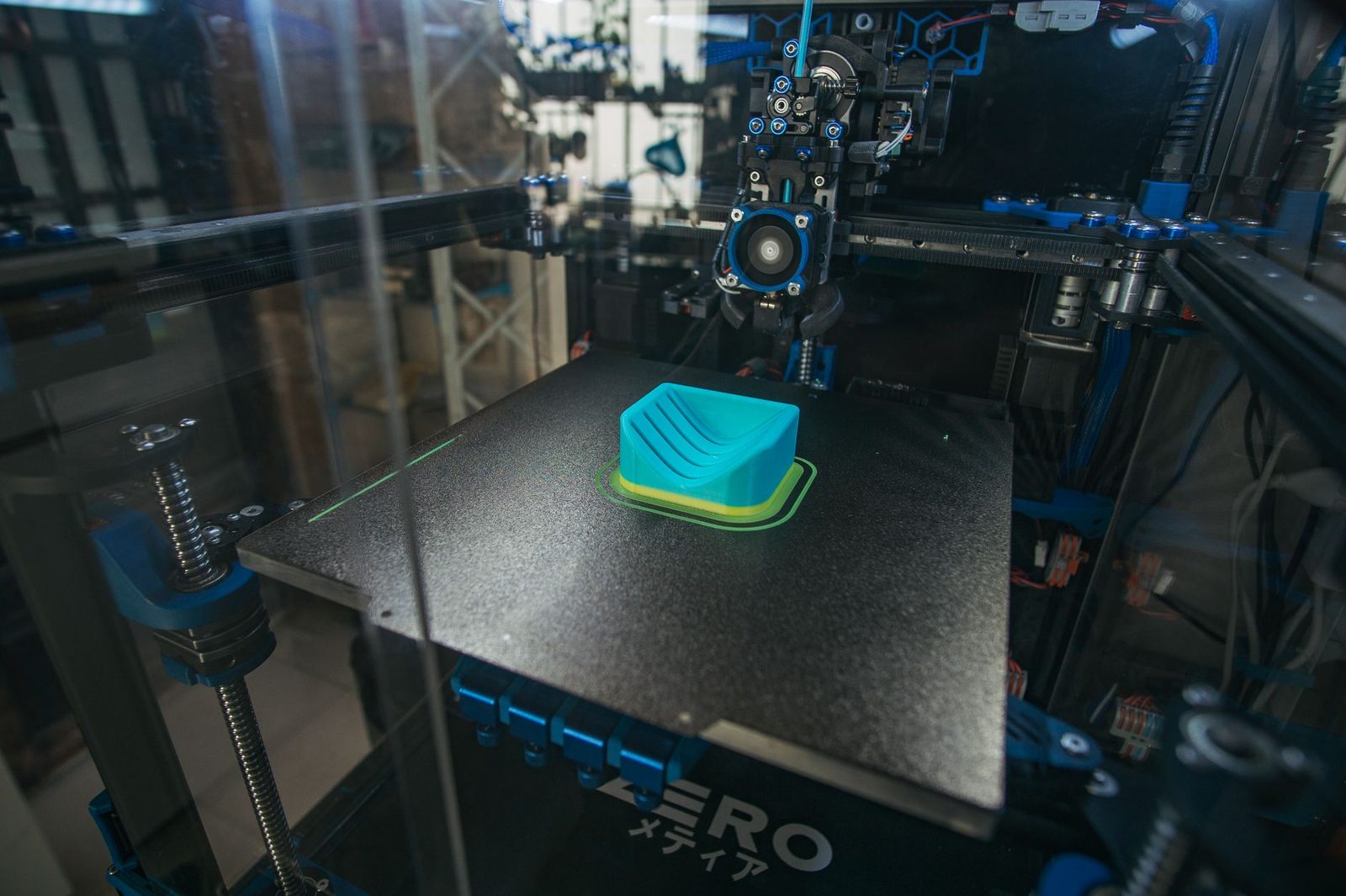 Impressora 3D fechada vista pela porta de vidro, com o cabeçote parado sobre uma peça azul recém-impressa na mesa
