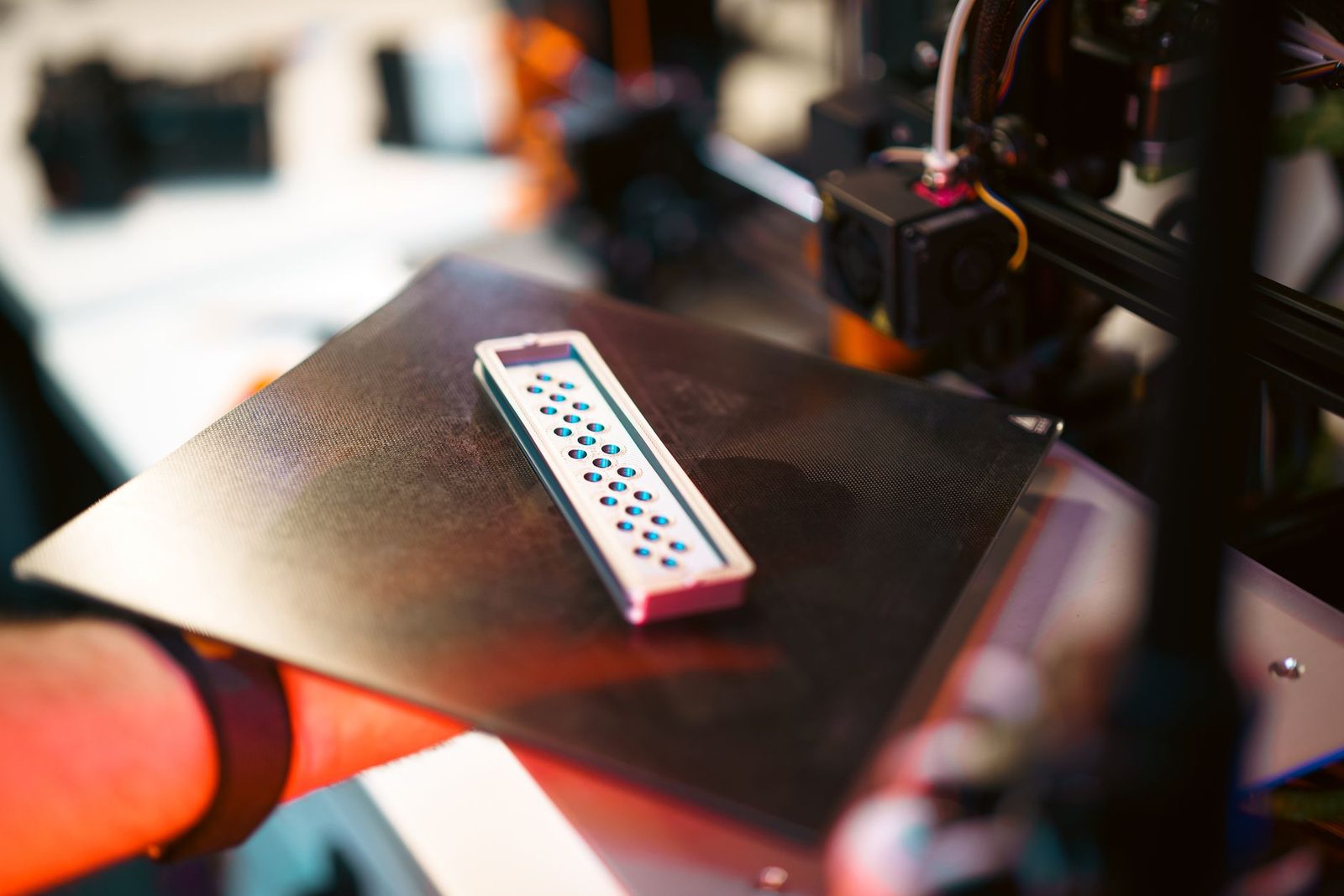 Mão segurando a placa de impressão com uma peça pequena recém-impressa, e o cabeçote da impressora 3D desfocado ao fundo