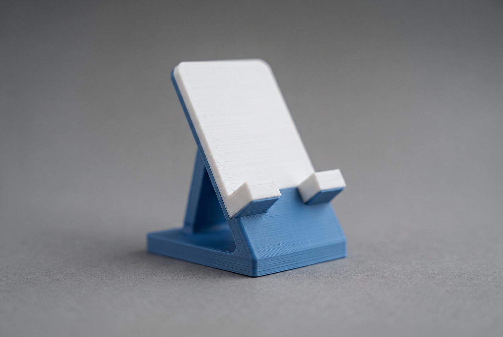 Suporte de celular impresso em 3D em duas cores, azul fosco e branco, com linhas de camada visíveis sobre fundo cinza liso