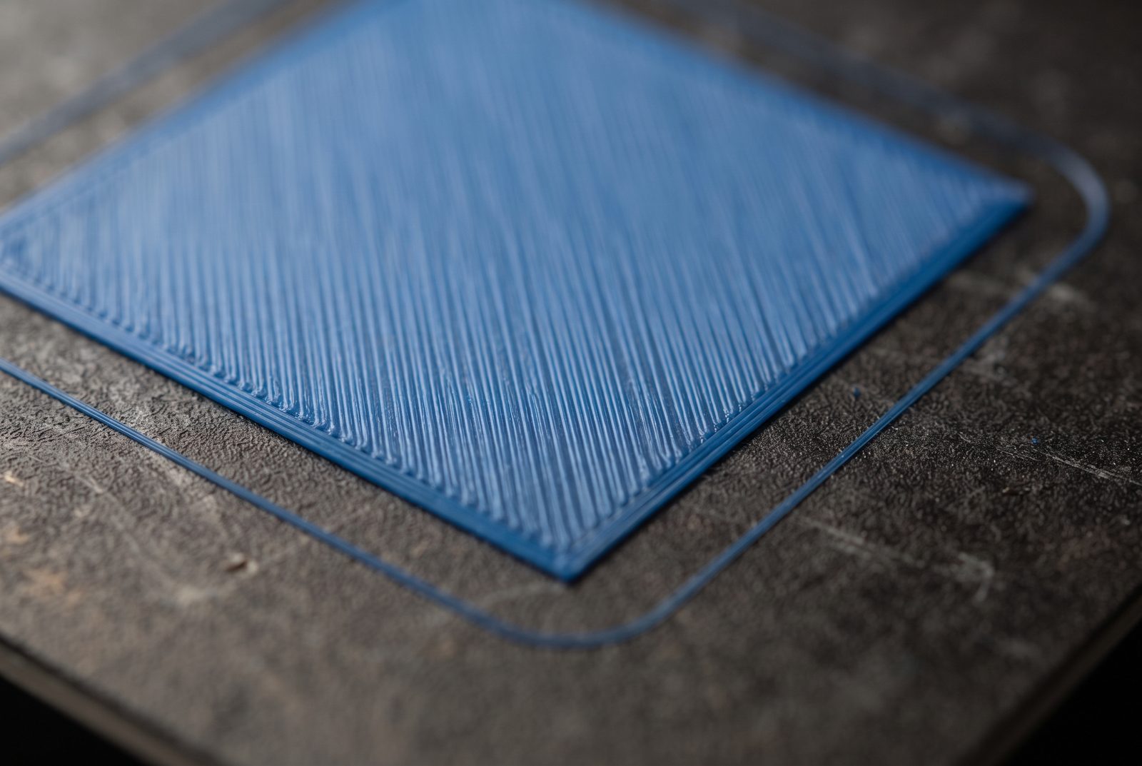 Primeira camada perfeitamente lisa de uma peça quadrada impressa em 3D em PLA azul sobre placa texturizada escura