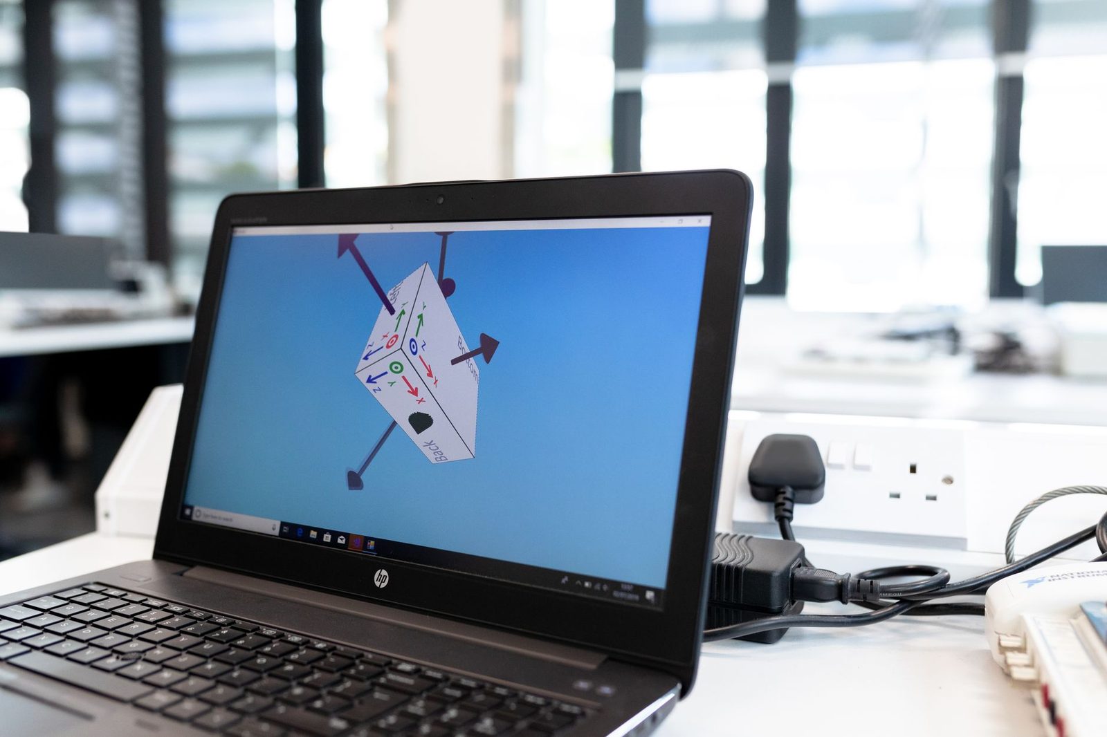 Notebook aberto numa mesa de escritório mostrando um bloco 3D com setas de eixo num programa de modelagem