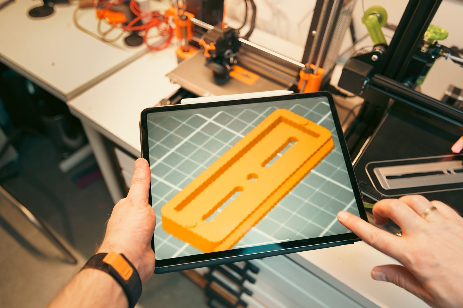 Mãos segurando um tablet com uma peça laranja em 3D na tela, diante de impressoras 3D numa oficina