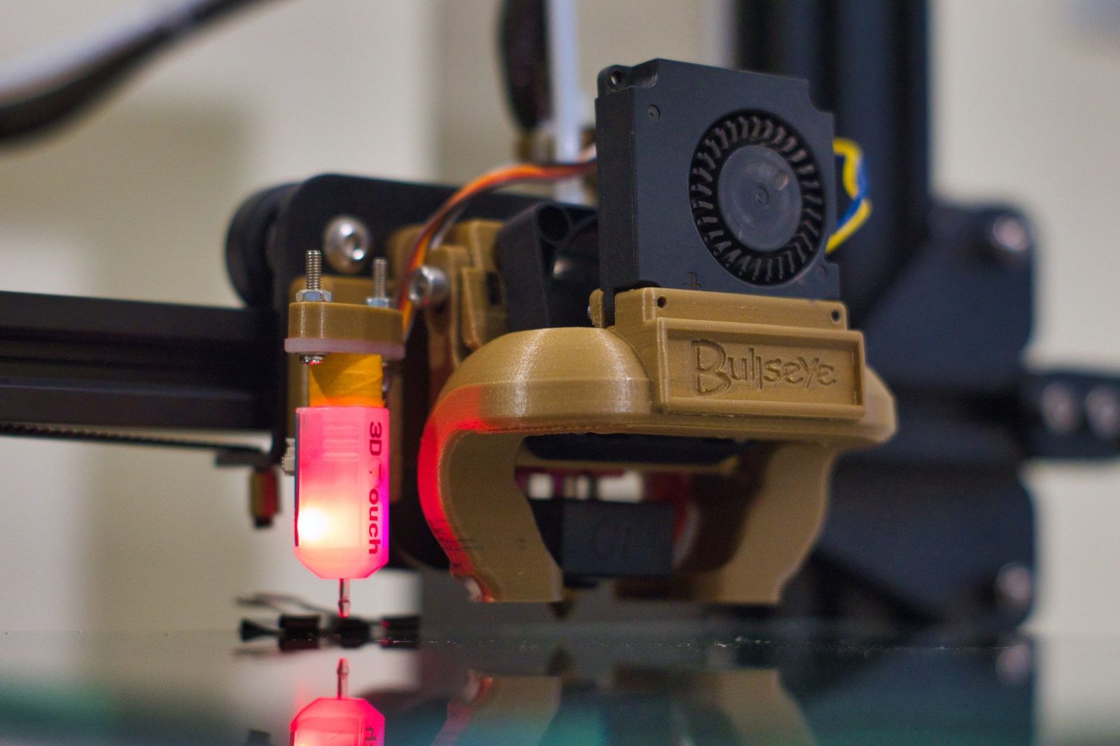 Cabeçote de impressora 3D visto de perto, com sensor de nivelamento aceso em vermelho e duto de ventilação impresso em filamento dourado
