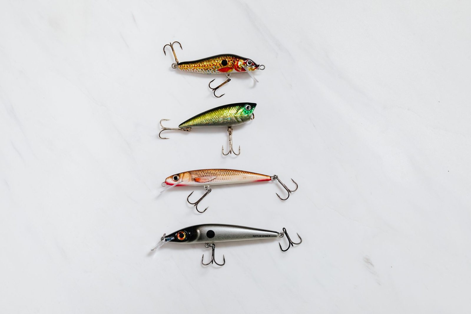 Quatro iscas artificiais duras pintadas imitando peixinhos, com garateias, enfileiradas sobre um tampo de mármore branco
