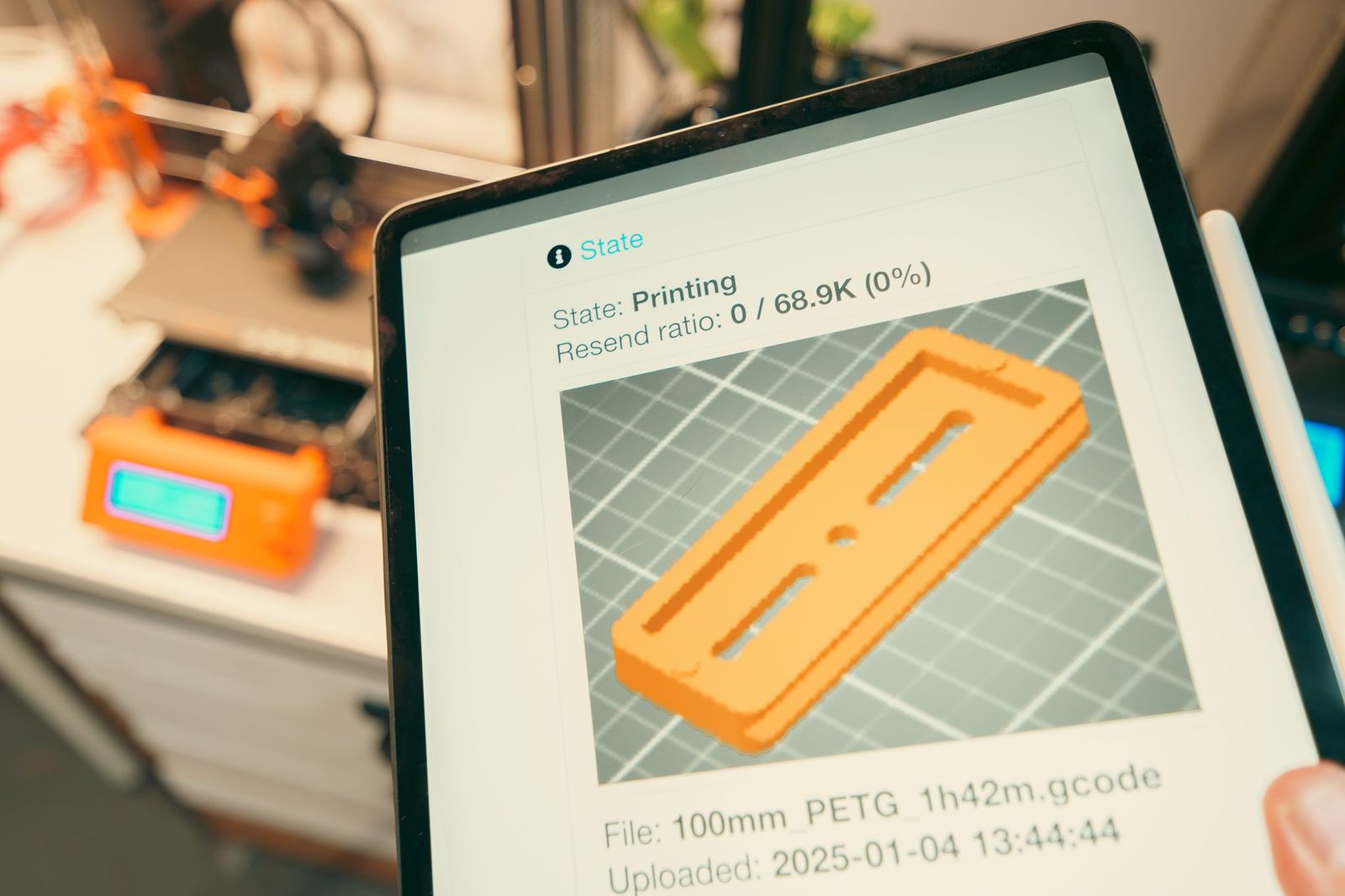 Tablet mostrando o estado de uma impressão em andamento, com uma impressora 3D desfocada ao fundo