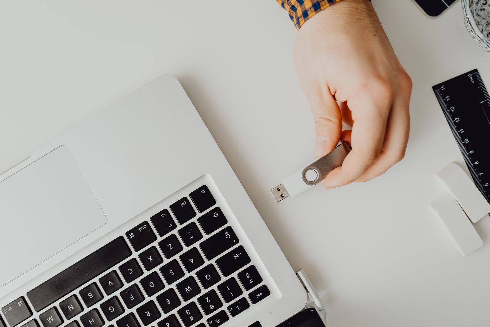 Mão segurando um pendrive branco ao lado de um notebook, prestes a encaixar na porta USB