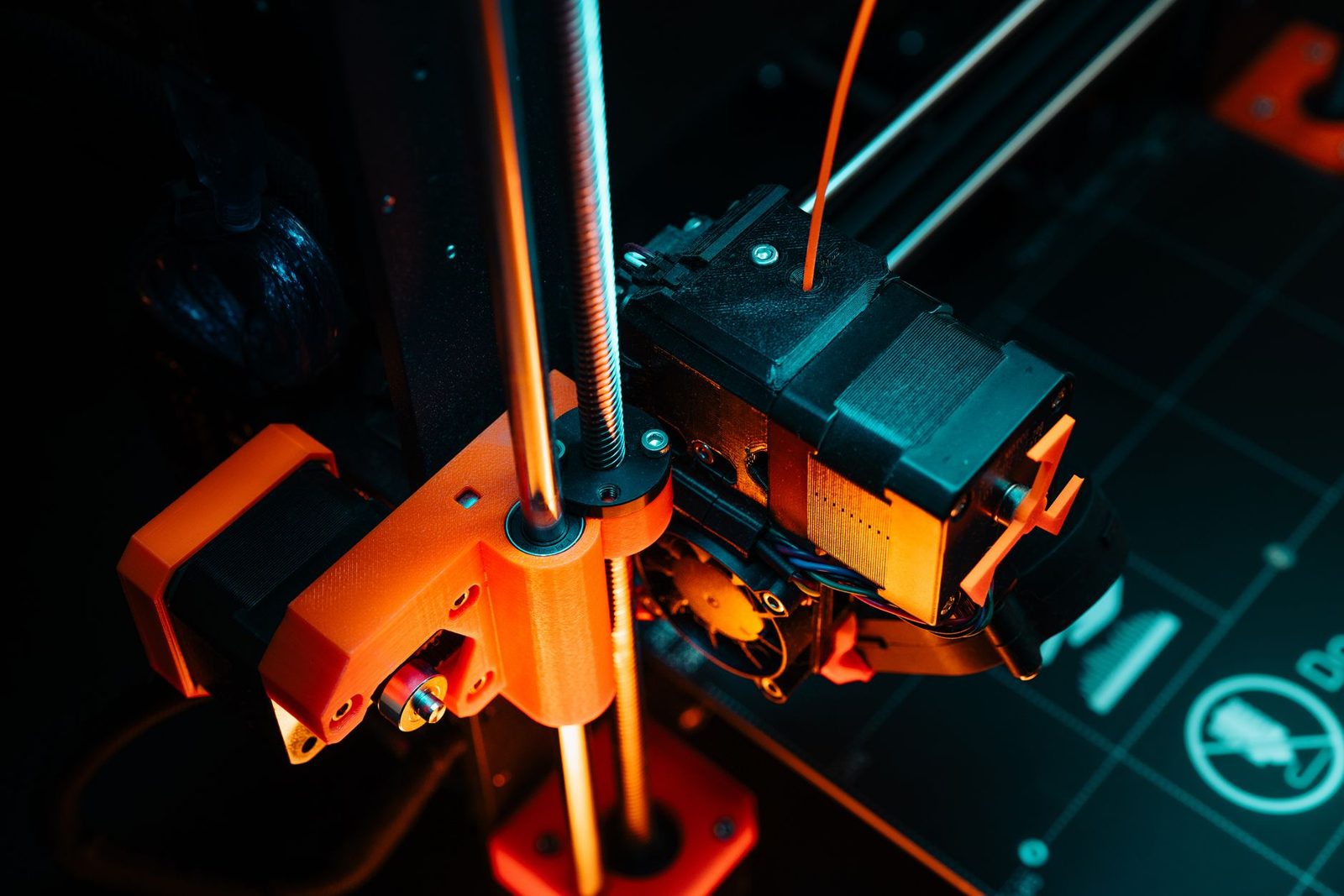 Extrusora de uma impressora 3D vista de cima, com o filamento laranja entrando pelo furo de alimentação