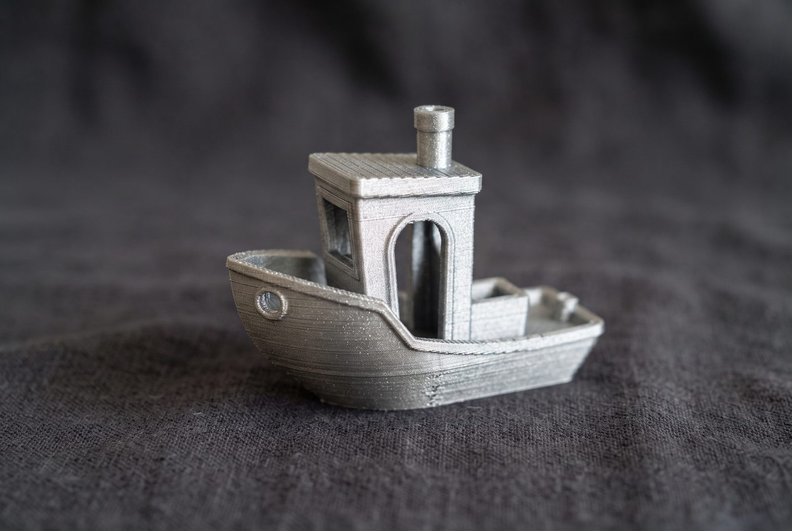 Barquinho Benchy impresso em 3D em PLA prateado metálico sobre tecido escuro, com linhas de camada visíveis
