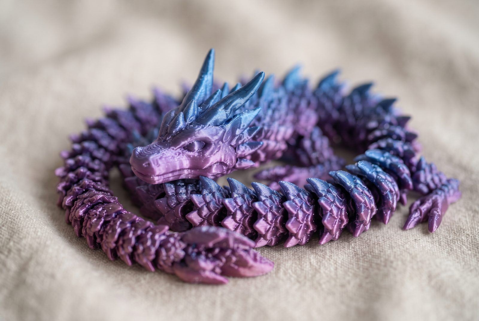 Dragão articulado impresso em 3D em PLA silk degradê roxo e azul enrolado sobre um tecido neutro