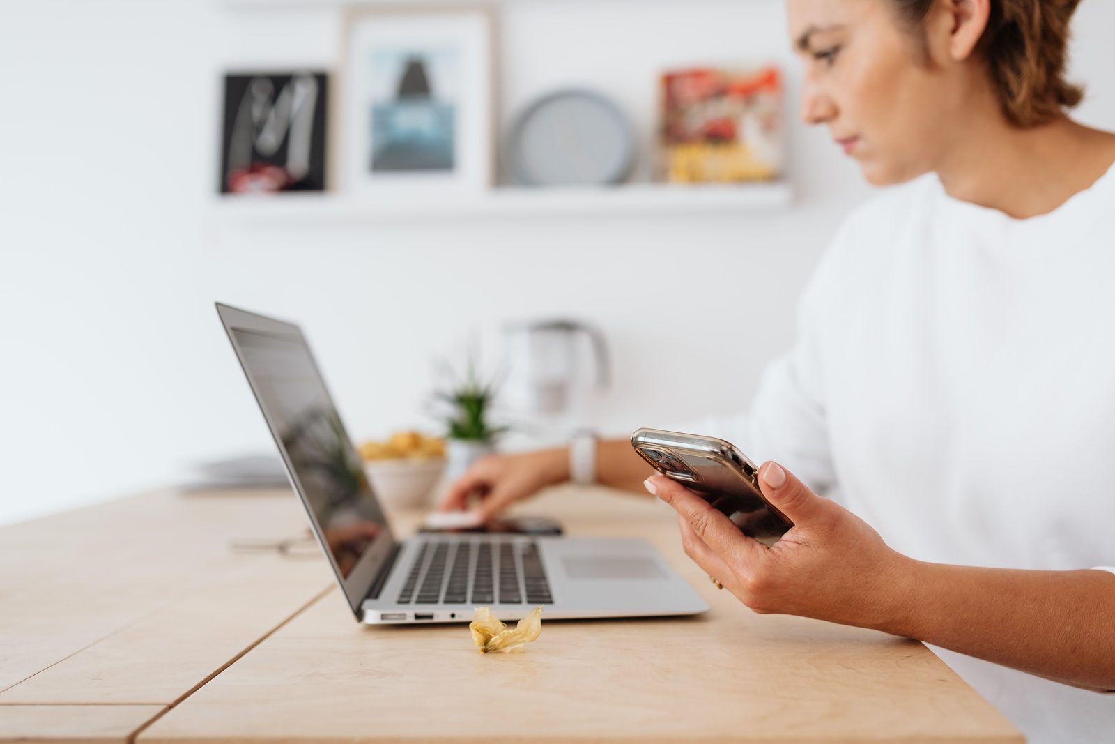 Mulher sentada a uma mesa de madeira olhando o celular, com o notebook aberto ao lado