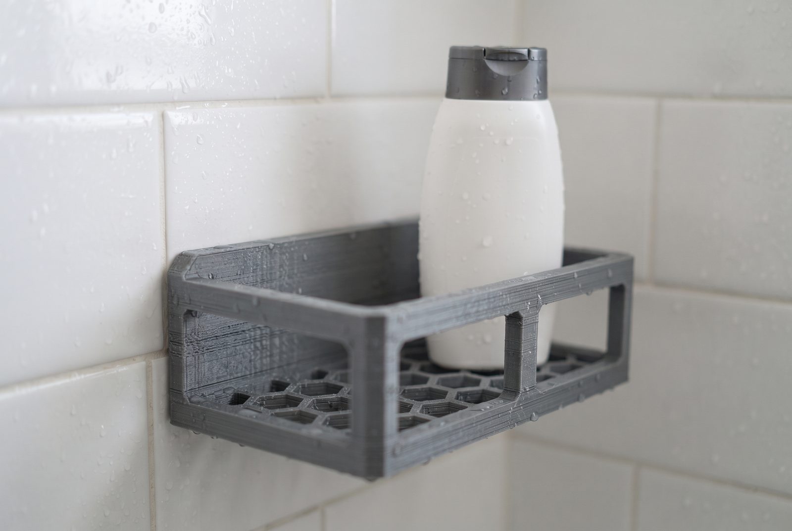 Prateleira de box impressa em 3D em PETG cinza segurando um frasco de shampoo sem rótulo, com gotas d'água no azulejo atrás