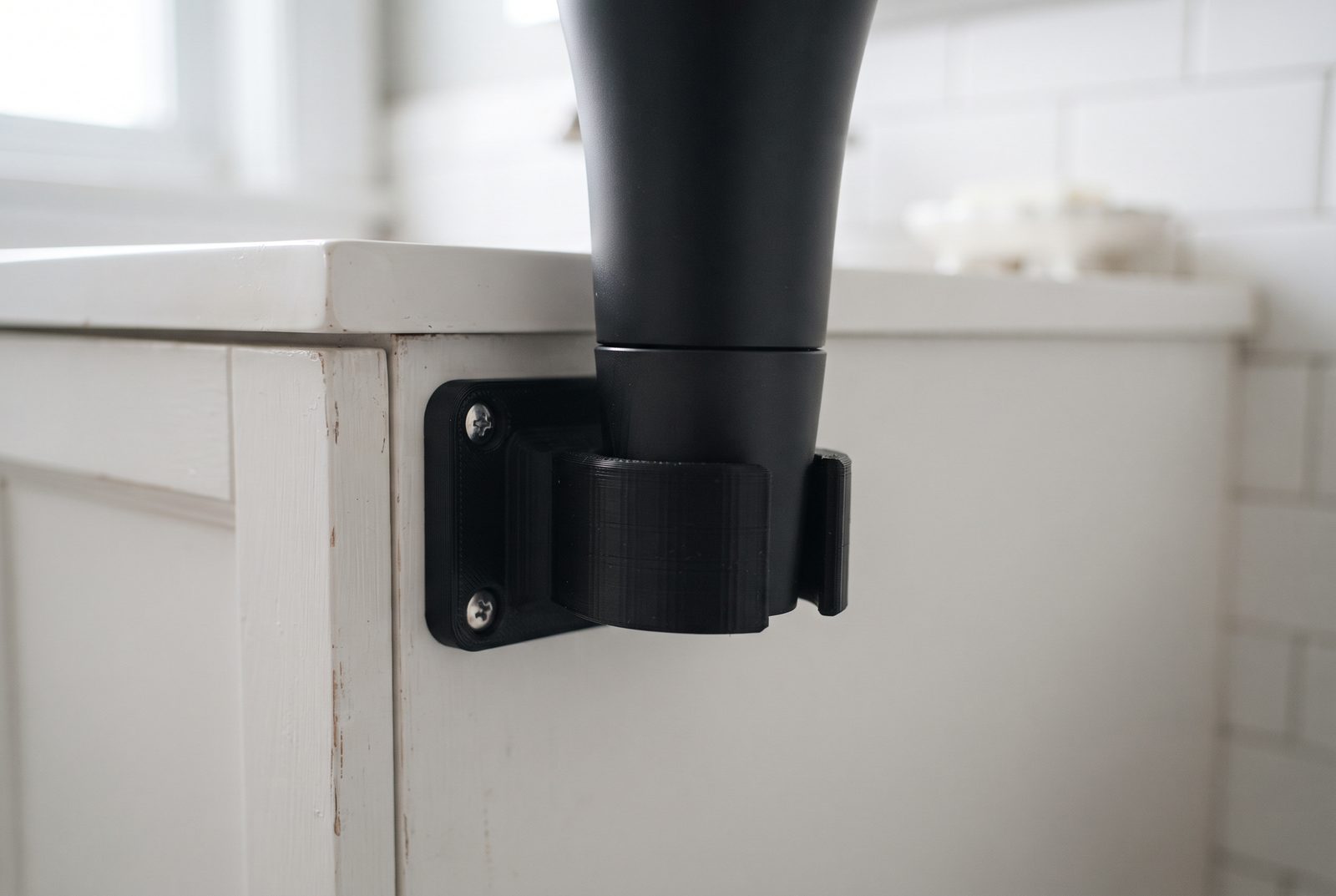 Suporte de secador de cabelo impresso em 3D em PLA preto fixado ao lado de um armário de banheiro liso, com o secador apoiado