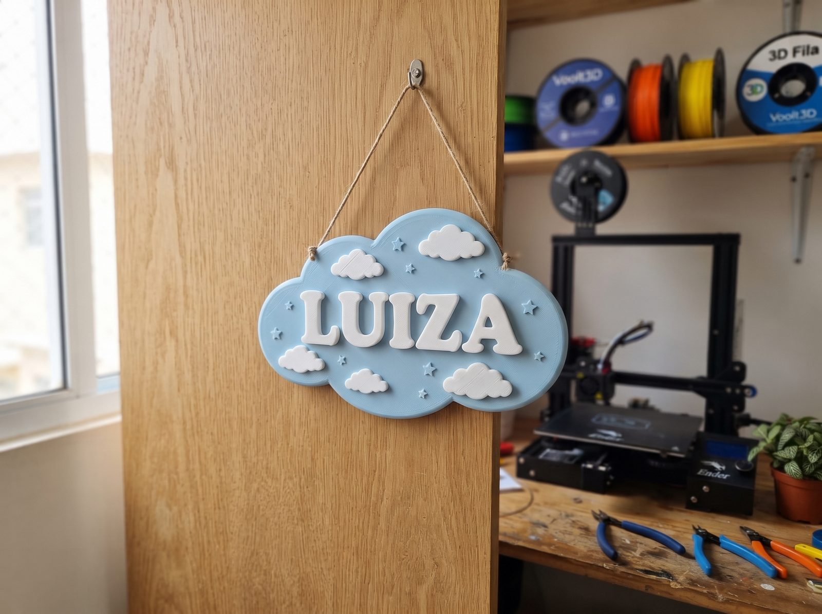 Placa de porta impressa em 3D em PLA azul pastel e branco com o nome do bebê em relevo e nuvens