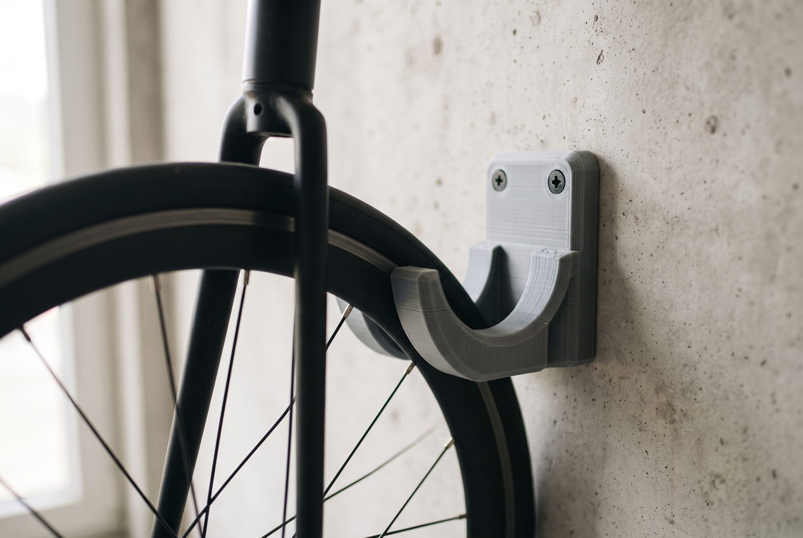 Gancho de parede impresso em 3D em PETG cinza apoiando a roda dianteira de uma bicicleta, parafusado numa parede de concreto