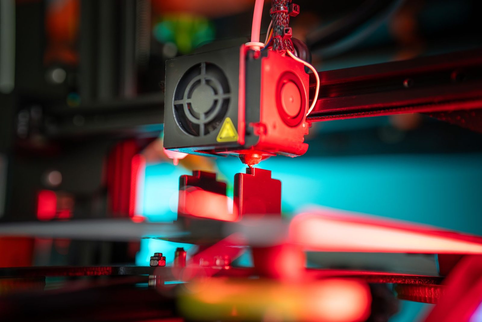 Bico de uma impressora 3D depositando filamento sobre uma peça de cantos retos, com a cena iluminada por luz vermelha e azul
