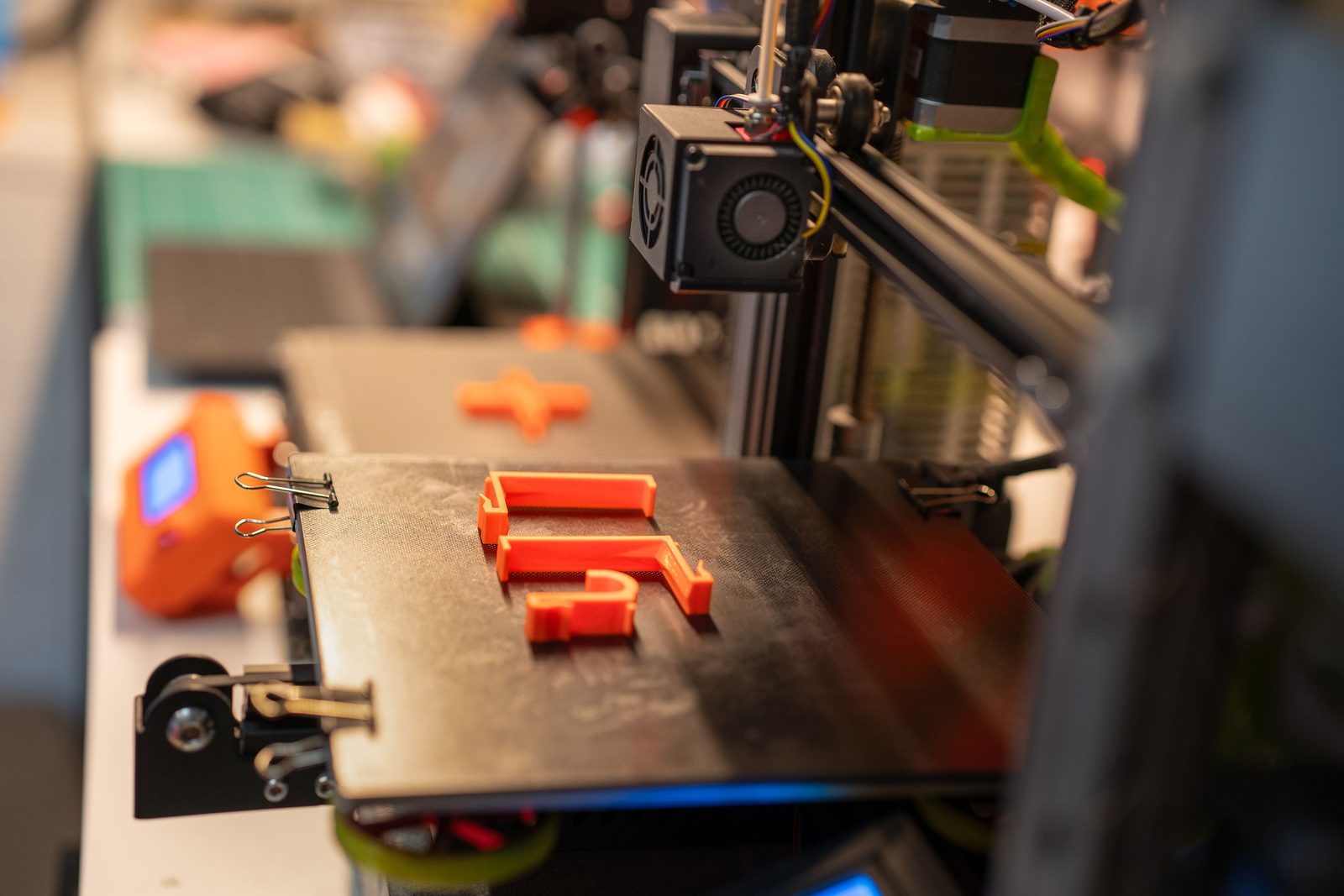 Três peças laranja finas, em formato de gancho, sobre a mesa preta de uma impressora 3D