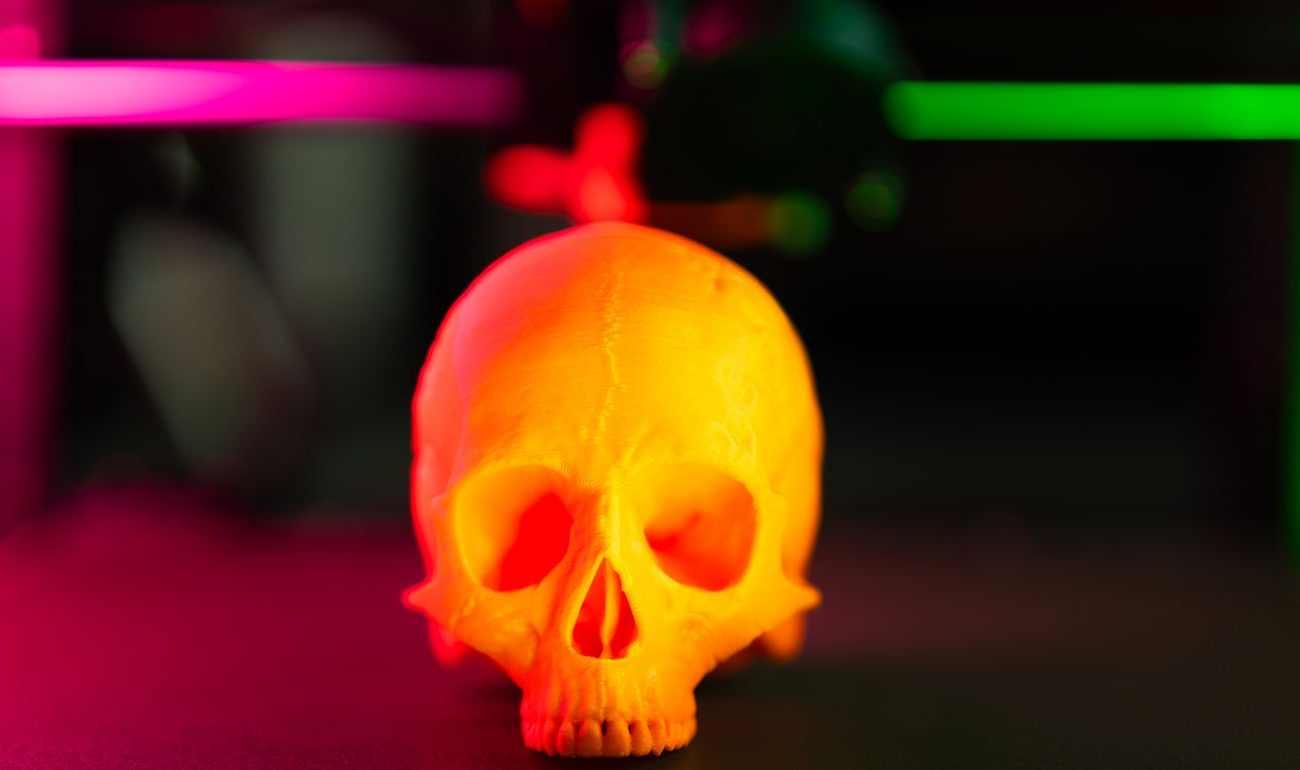 Caveira impressa em 3D em PLA laranja sobre a mesa de uma impressora, com luz rosa e verde ao fundo
